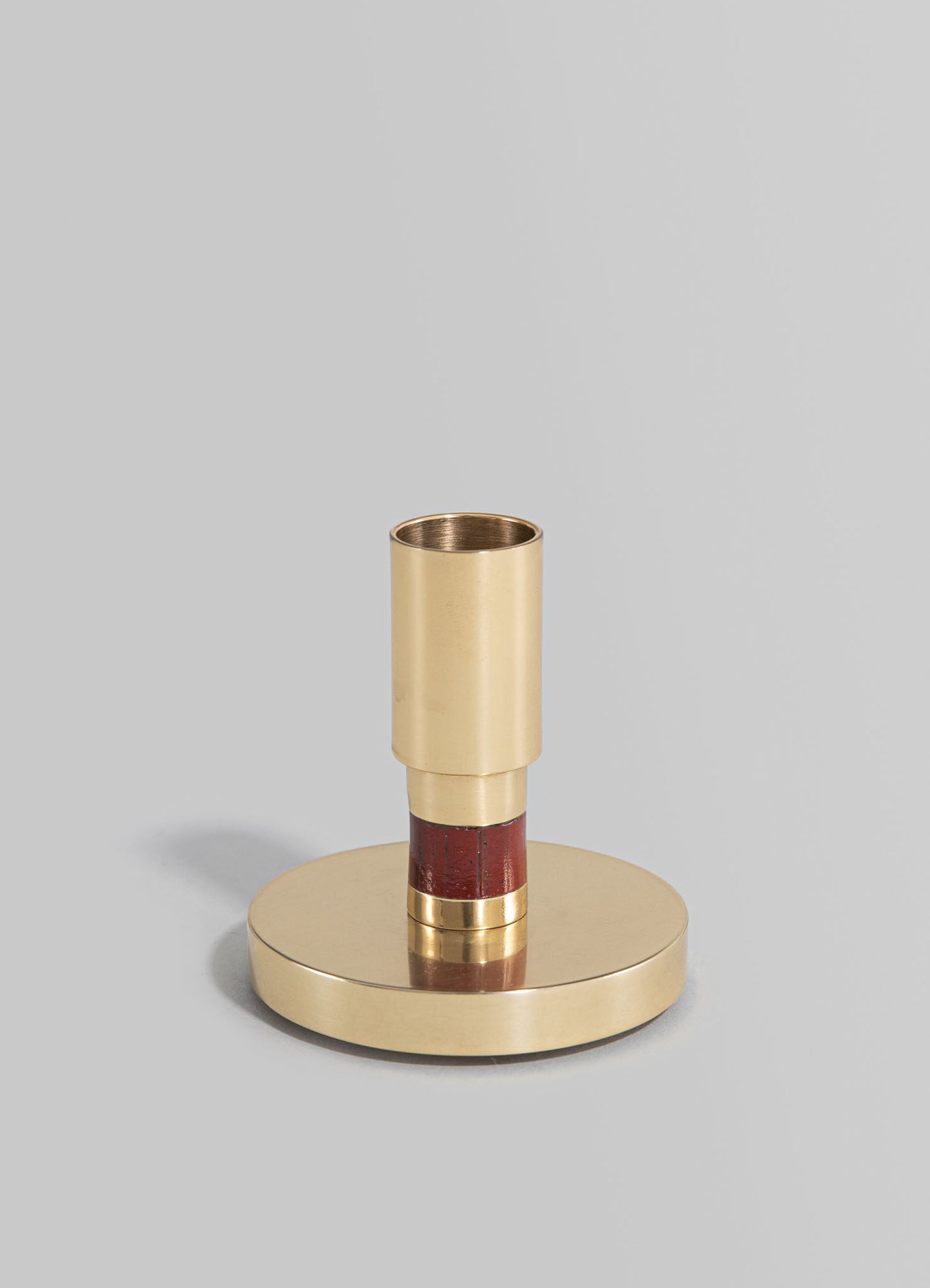 A modern brass candle holder with a cylindrical cup and a round base, featuring a red accent band around the stem. The finish is polished and metallic, with a minimalist and contemporary style.