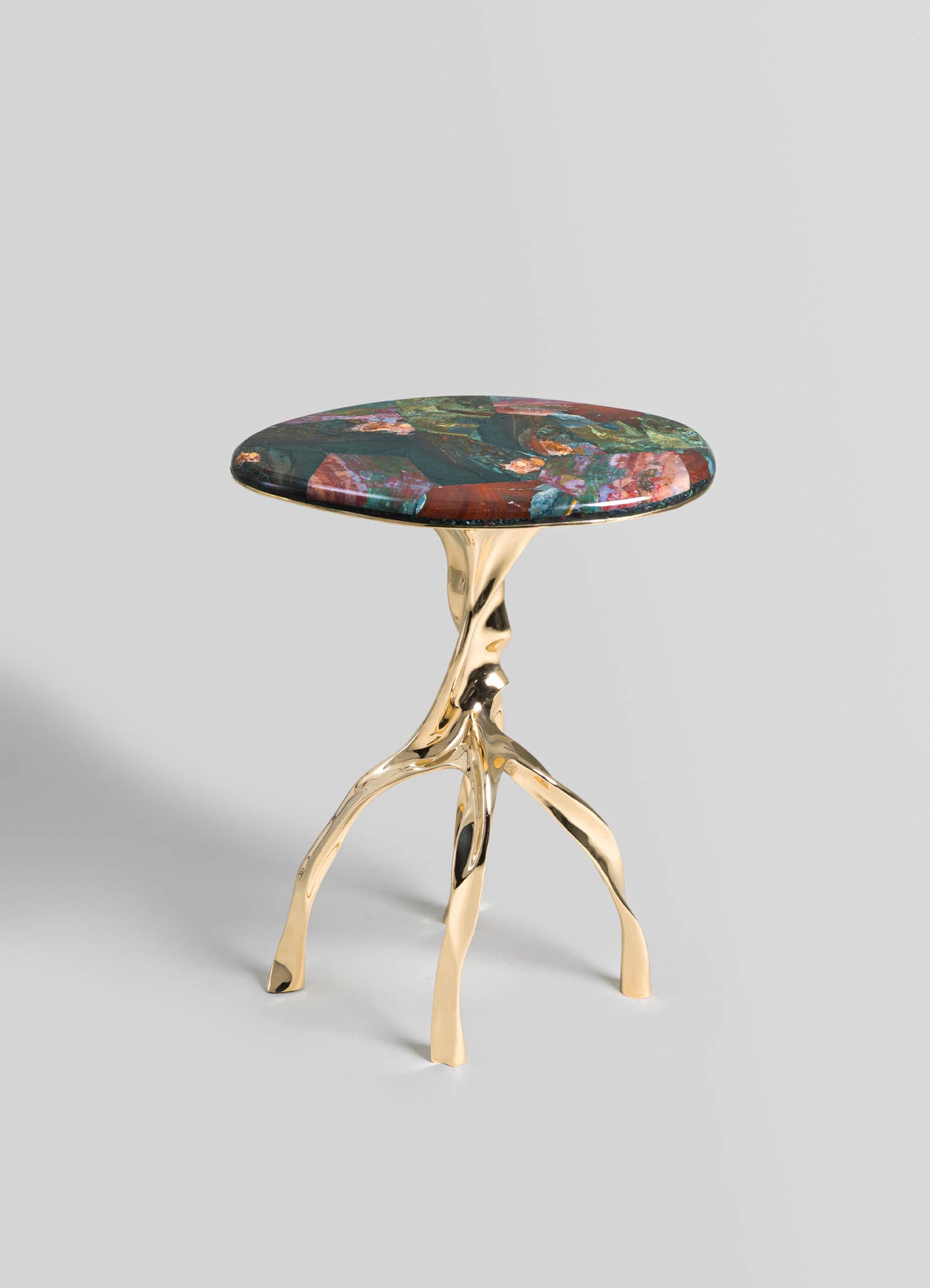 A small side table with a round top featuring a colorful, abstract pattern in shades of red, pink, green, and blue, paired with a sculptural gold metal base with three curved legs.