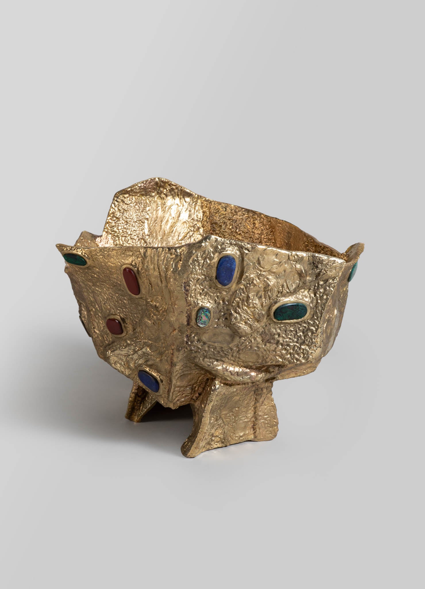 A decorative bowl made of textured gold-toned metal with an irregular, organic shape and embedded oval gemstones in red, blue, green, and turquoise.