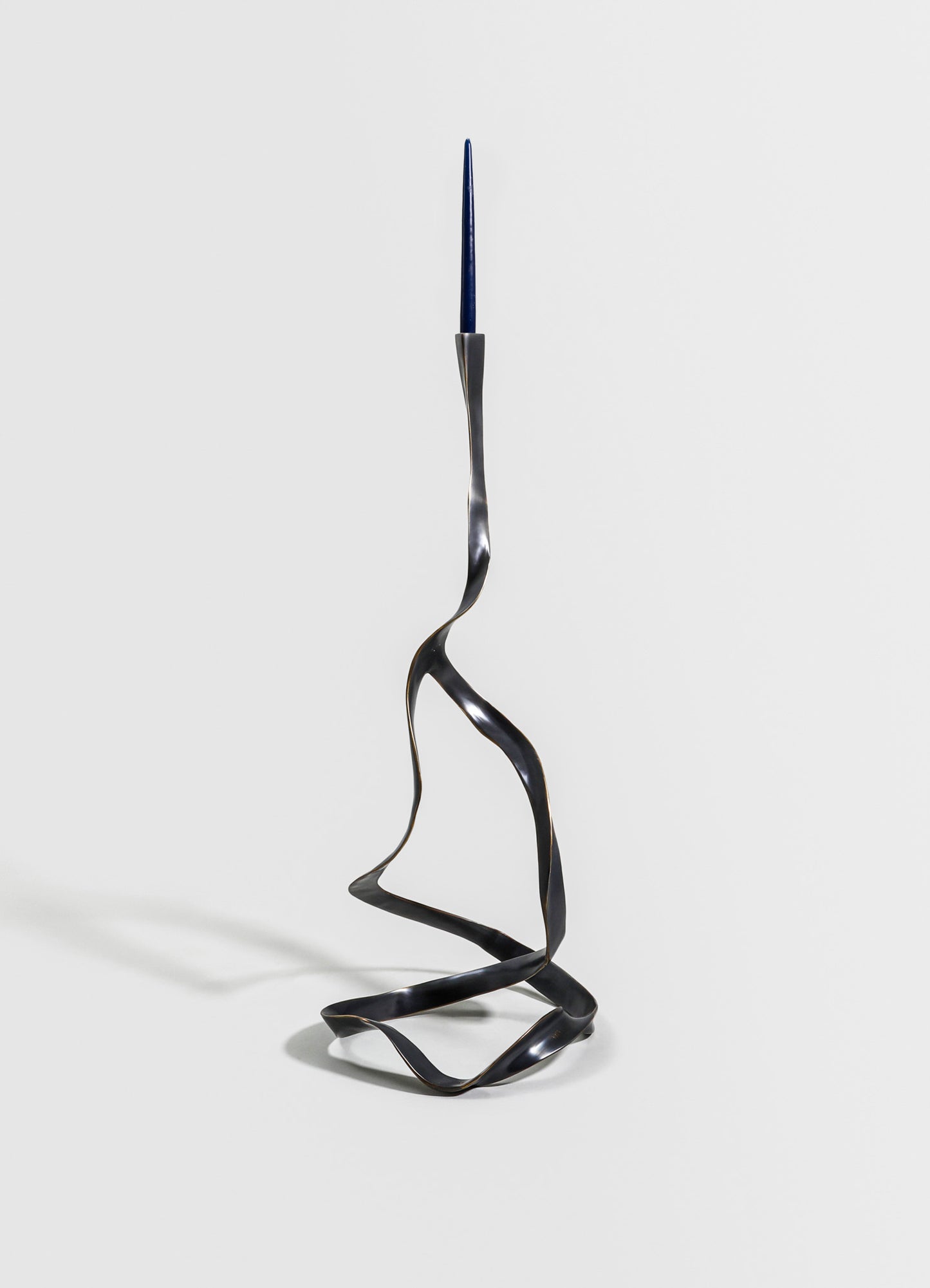 A sculptural candlestick holder made of sleek, anodized metal with a twisted, wavy design, supporting a single blue taper candle. The design features an abstract, contemporary style with a metallic finish against a plain background.