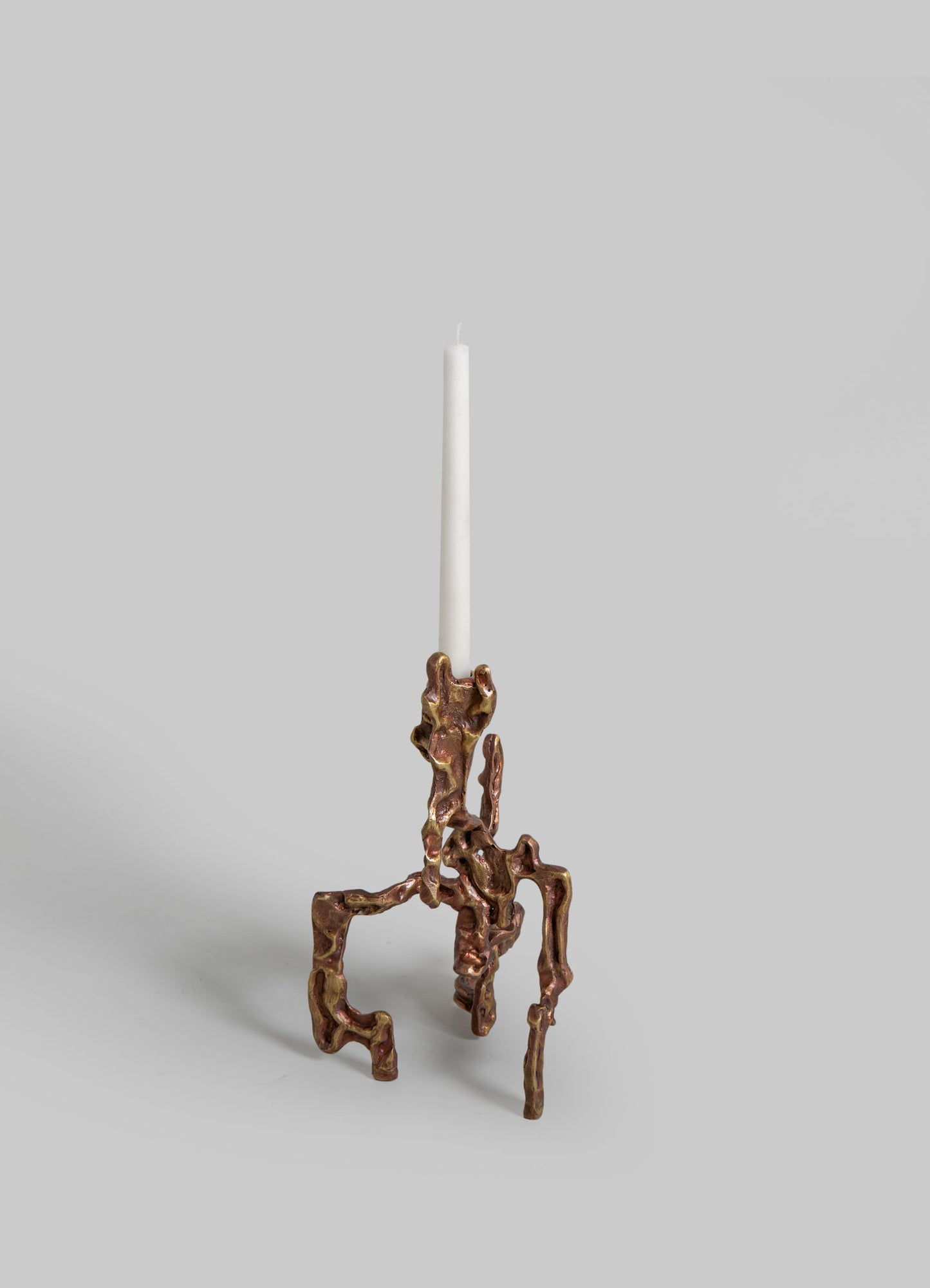 A decorative candle holder made of metal with an organic, irregular shape in a metallic bronze finish, holding a tall white taper candle. The design is abstract and sculptural, with a contemporary and artistic style.