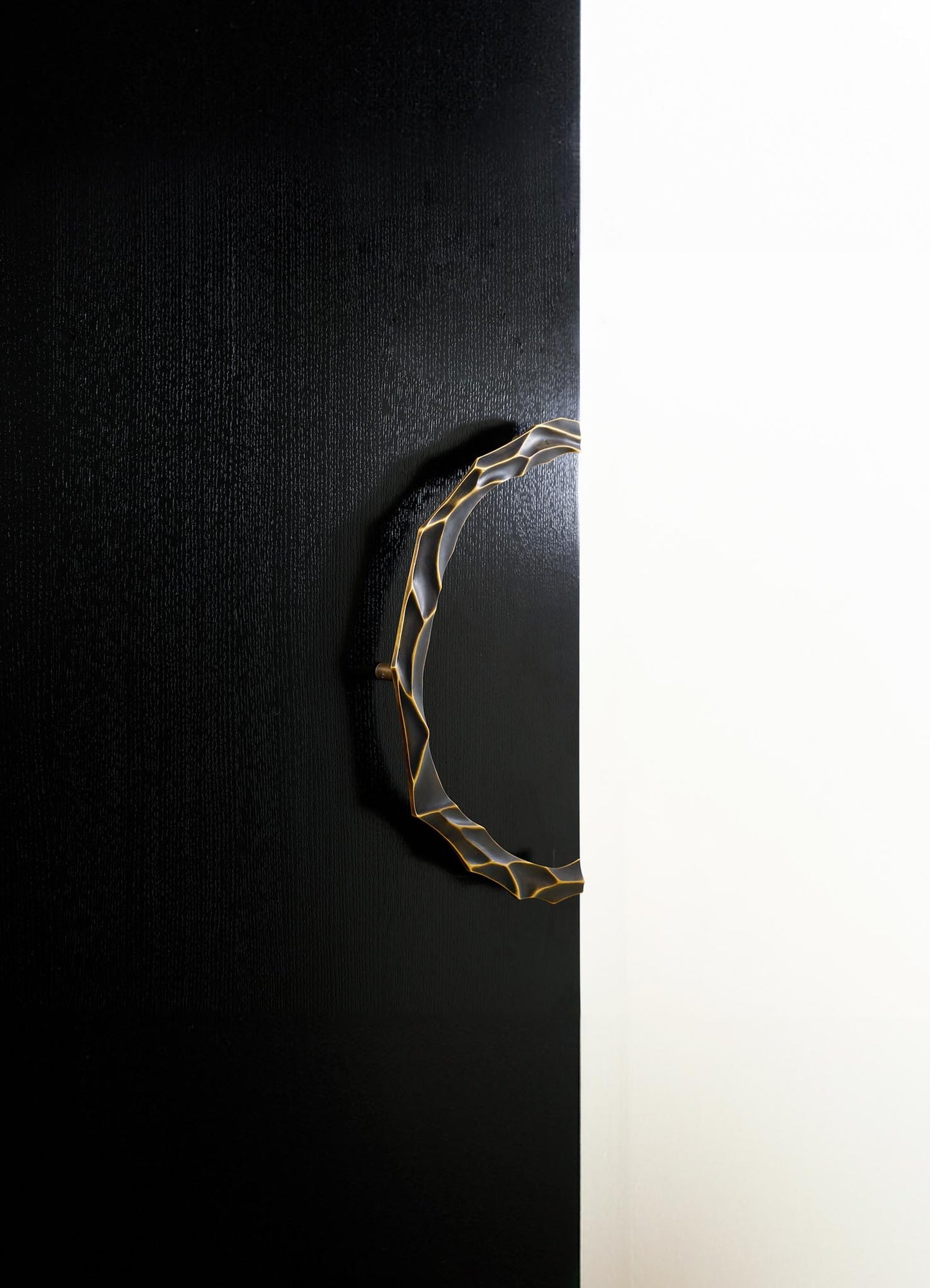 A modern wall-mounted light fixture with an irregular, organic-shaped metal frame finished in brass, emitting warm light against a black textured wall and a white background.