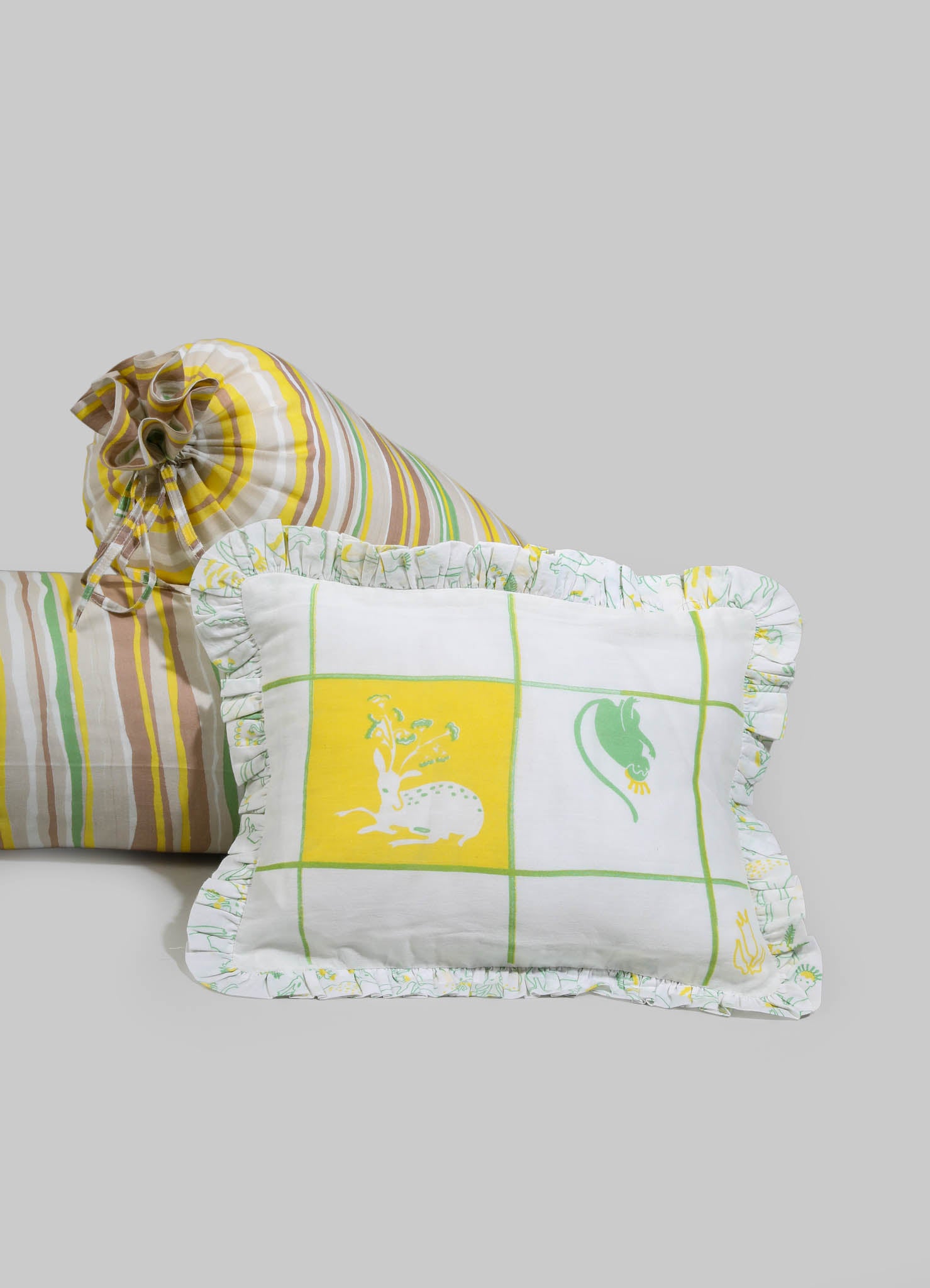 Fabric cushions with striped and printed patterns in shades of yellow, green, beige, and white, featuring ruffled edges. One cushion has a grid design with animal motifs, while another has a striped design with a drawstring closure.