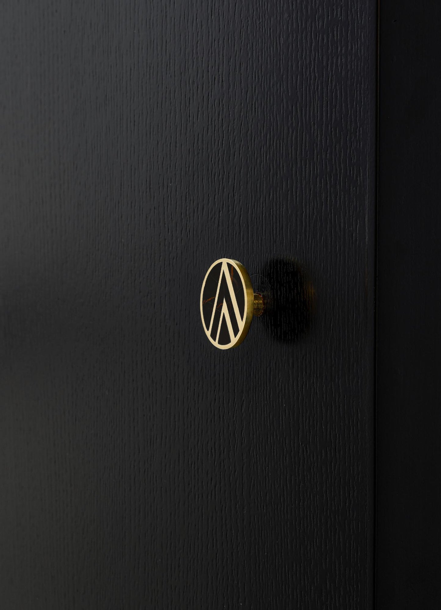 A black wooden wardrobe with a textured surface and a round brass handle featuring a geometric cutout pattern. The wardrobe design is minimalist and contemporary.