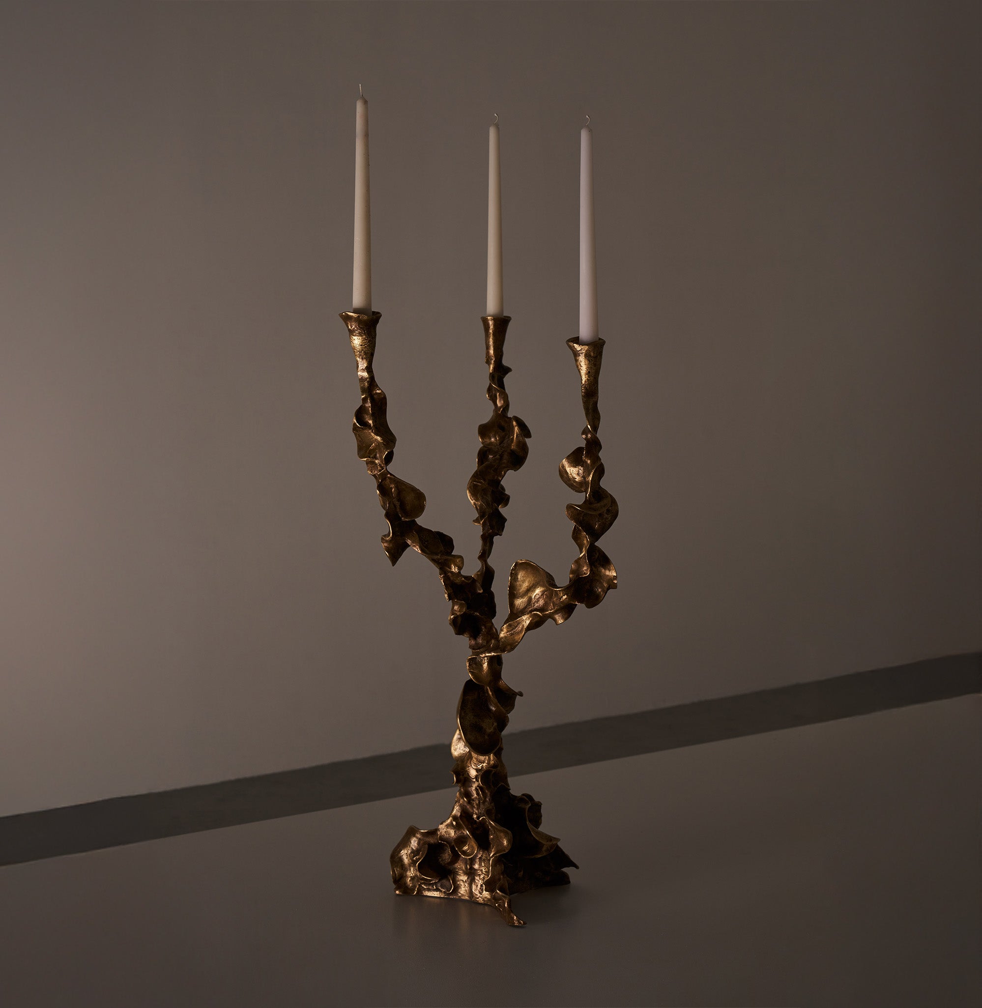 A bronze candle holder with an organic, sculptural design holding three white taper candles. The piece has a textured, irregular surface and a modern, artistic style.