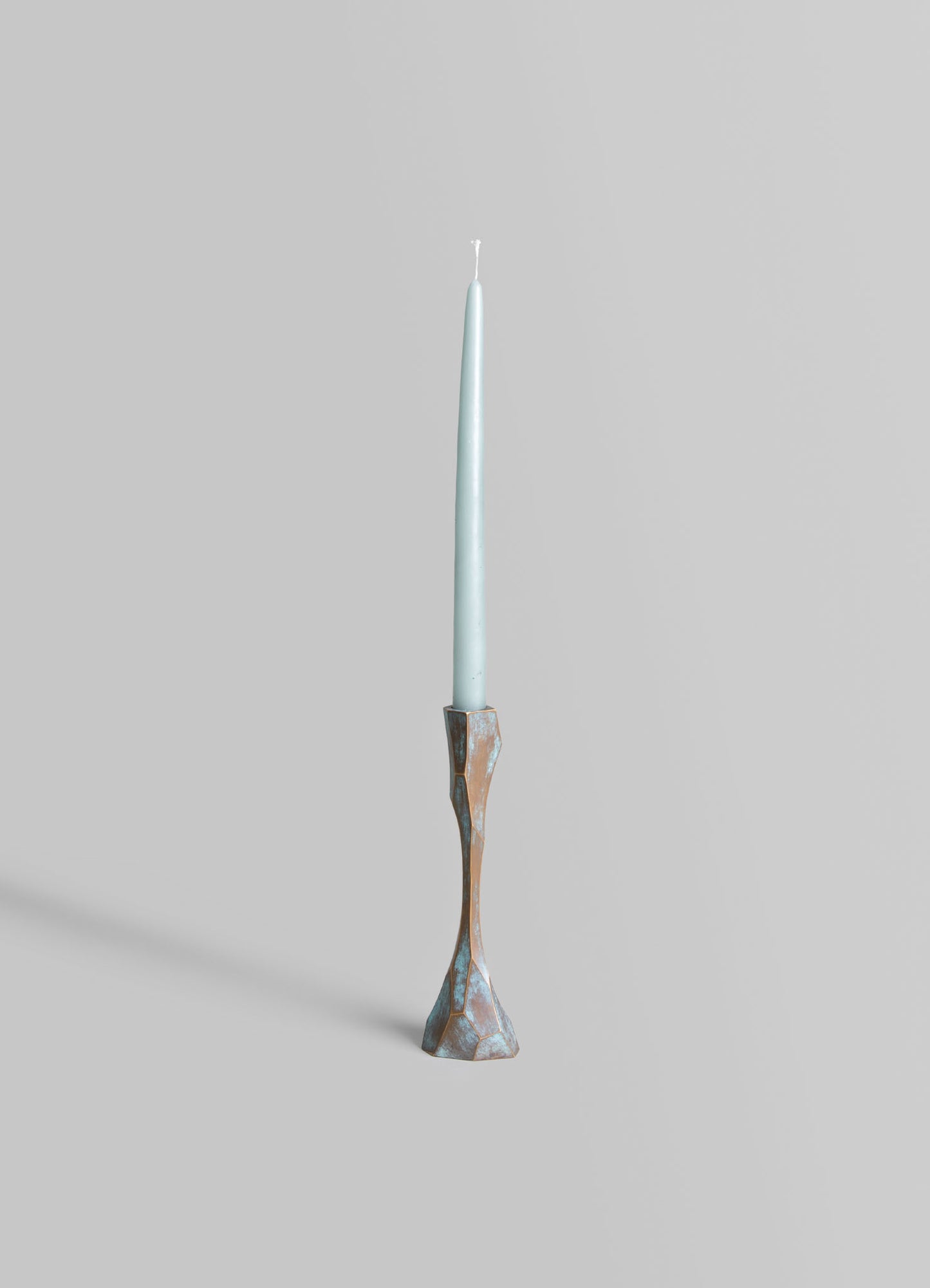 A tall candlestick holder made of metal with a rustic, textured finish in bronze and blue tones, featuring an elongated, sculptural form. It holds a slender, pale blue taper candle.