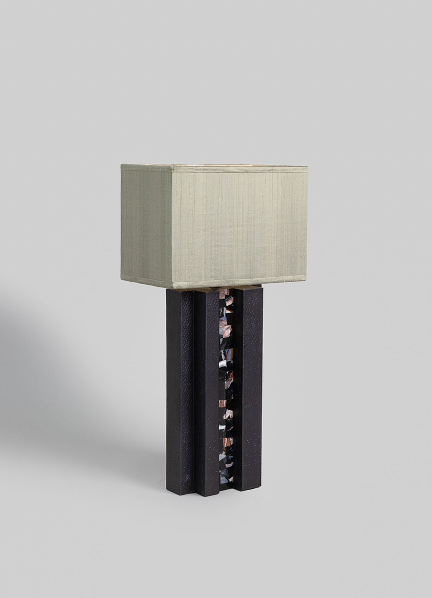 A table lamp featuring a rectangular fabric lampshade in light beige with subtle vertical texture, mounted on a dark wooden base with a central decorative element made of mosaic-style tiles.