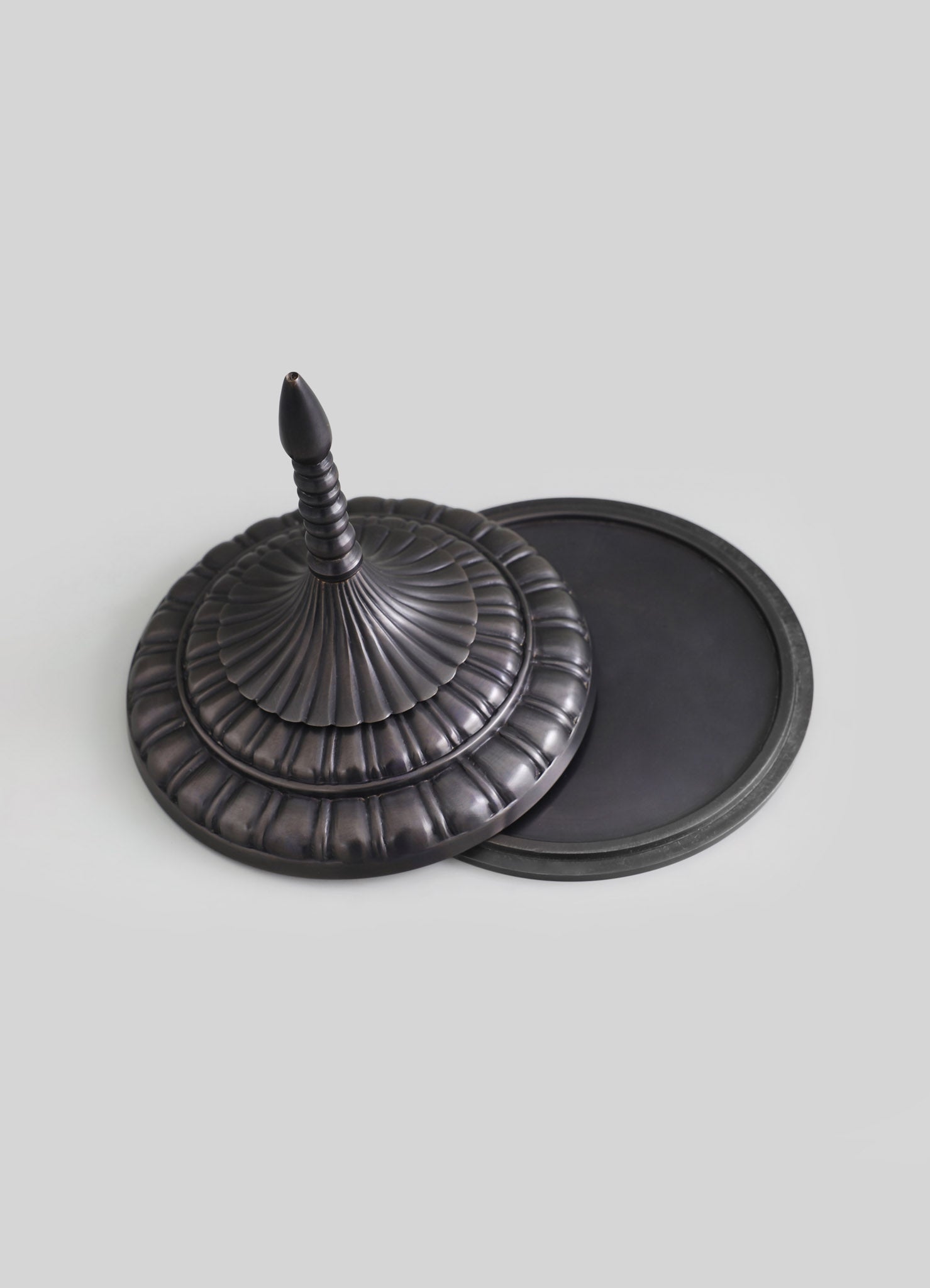 A dark metal, intricately designed incense holder with a fluted, circular base and a tall, pointed finial. The piece has a handcrafted, vintage style with detailed ridges and a matte finish.