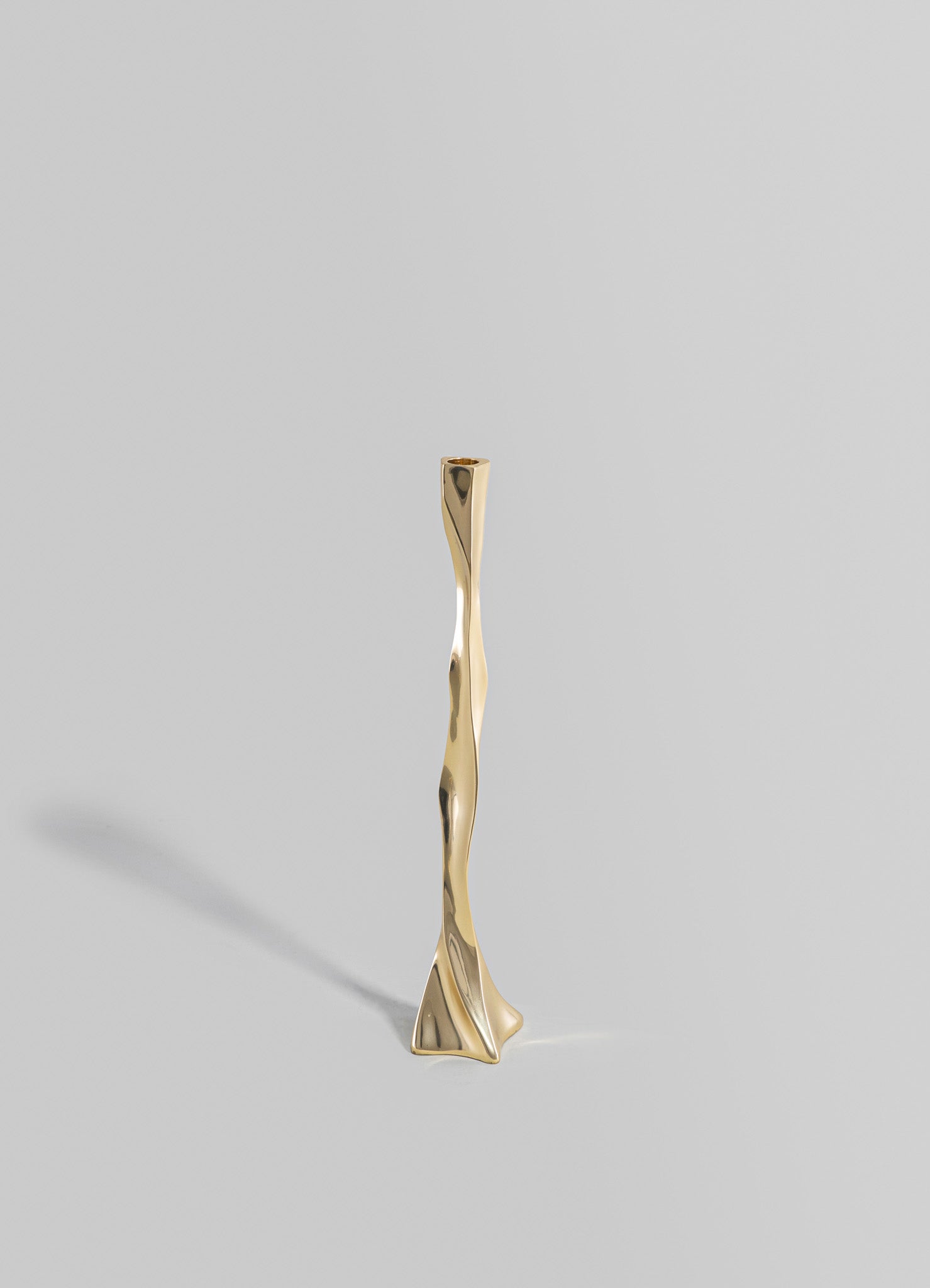 A sculptural candlestick holder made of polished brass with a wavy, organic form, featuring a slender vertical shape on a flat base.