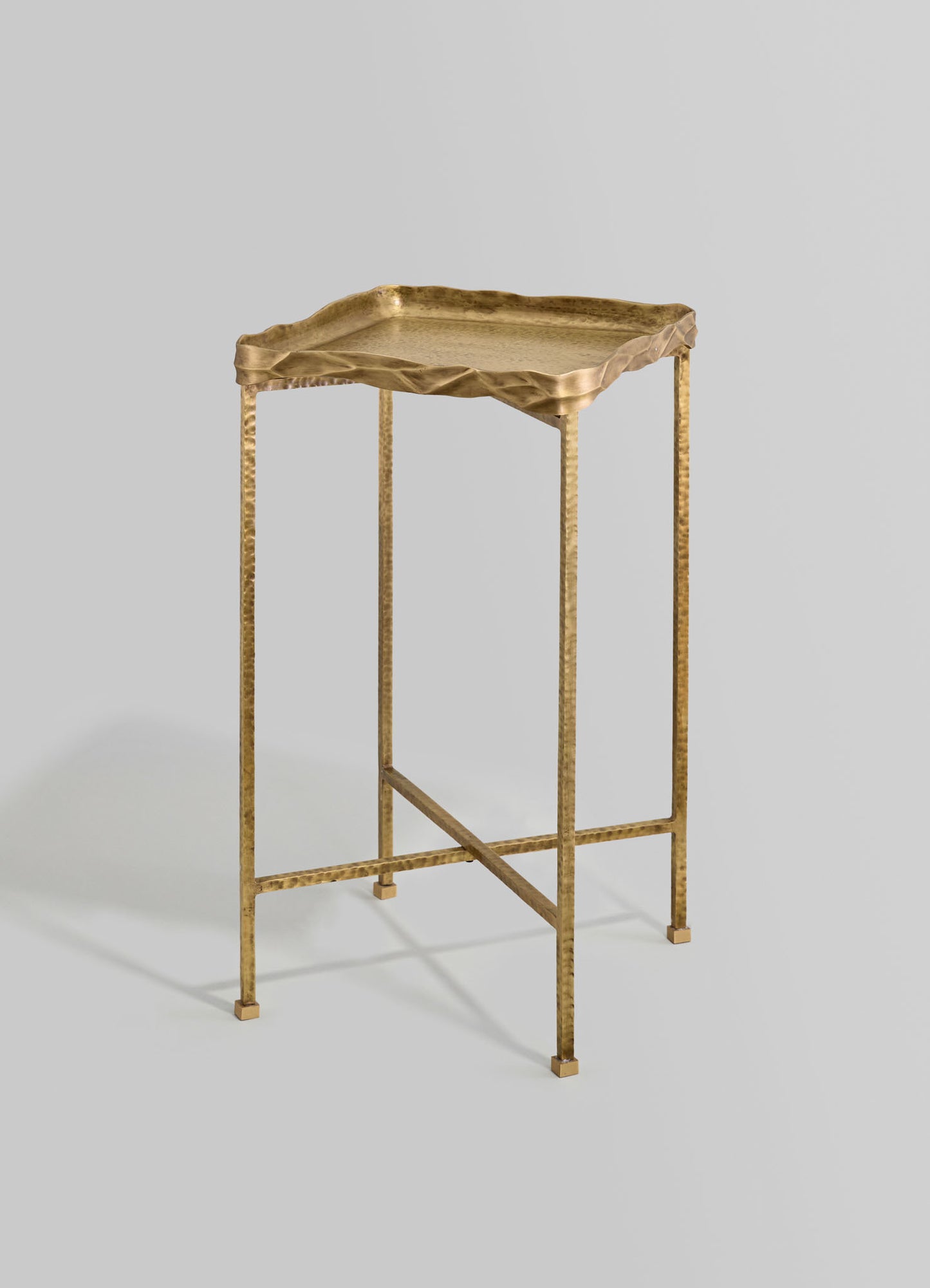 A gold-colored metal side table with a hammered finish, featuring a textured, irregular-edge tray top and slender, straight legs connected by a crossbar for support.