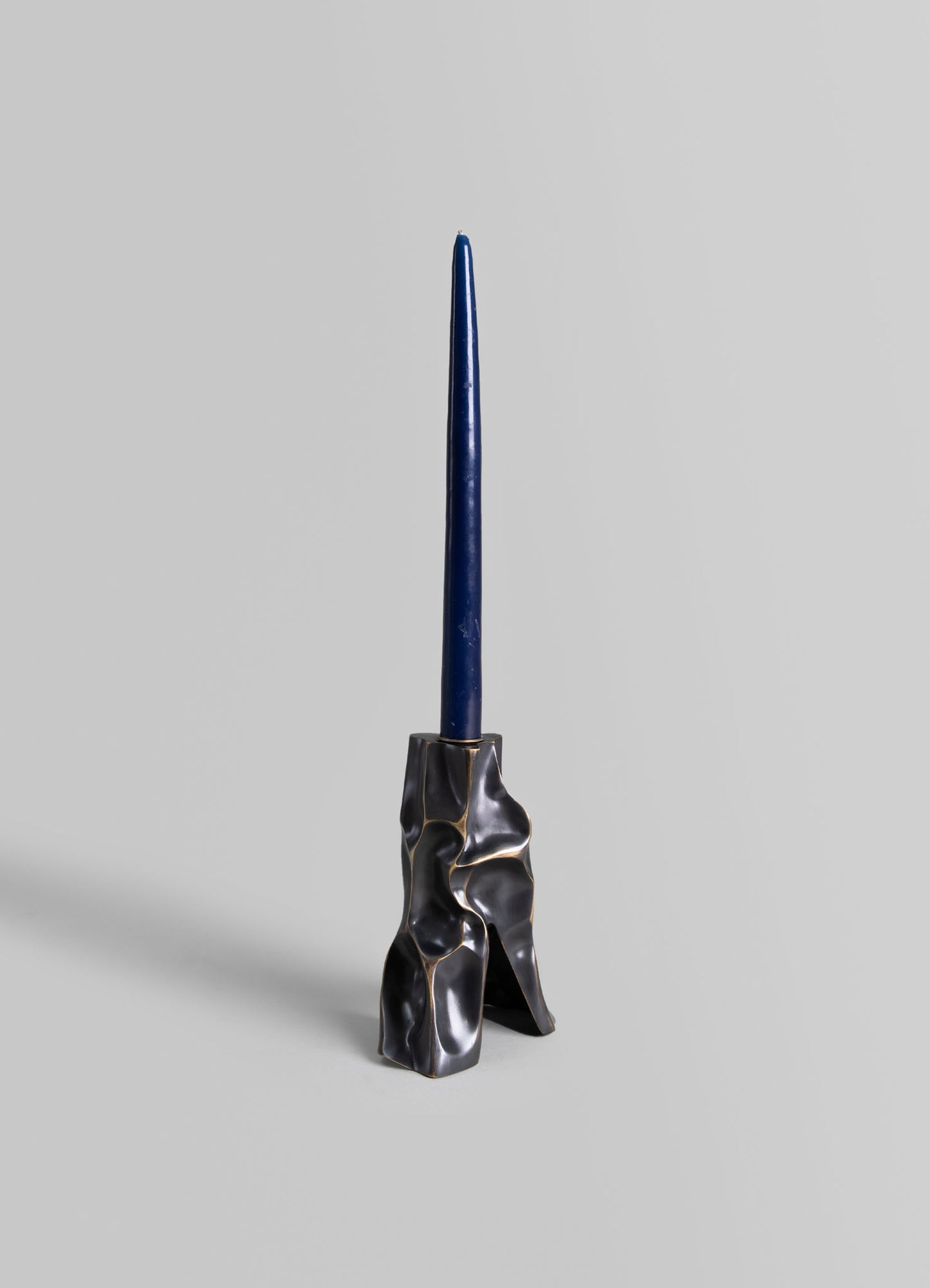 A tall, slender blue candle in a sculptural black and gold resin candle holder with an organic, irregular shape set against a plain background.