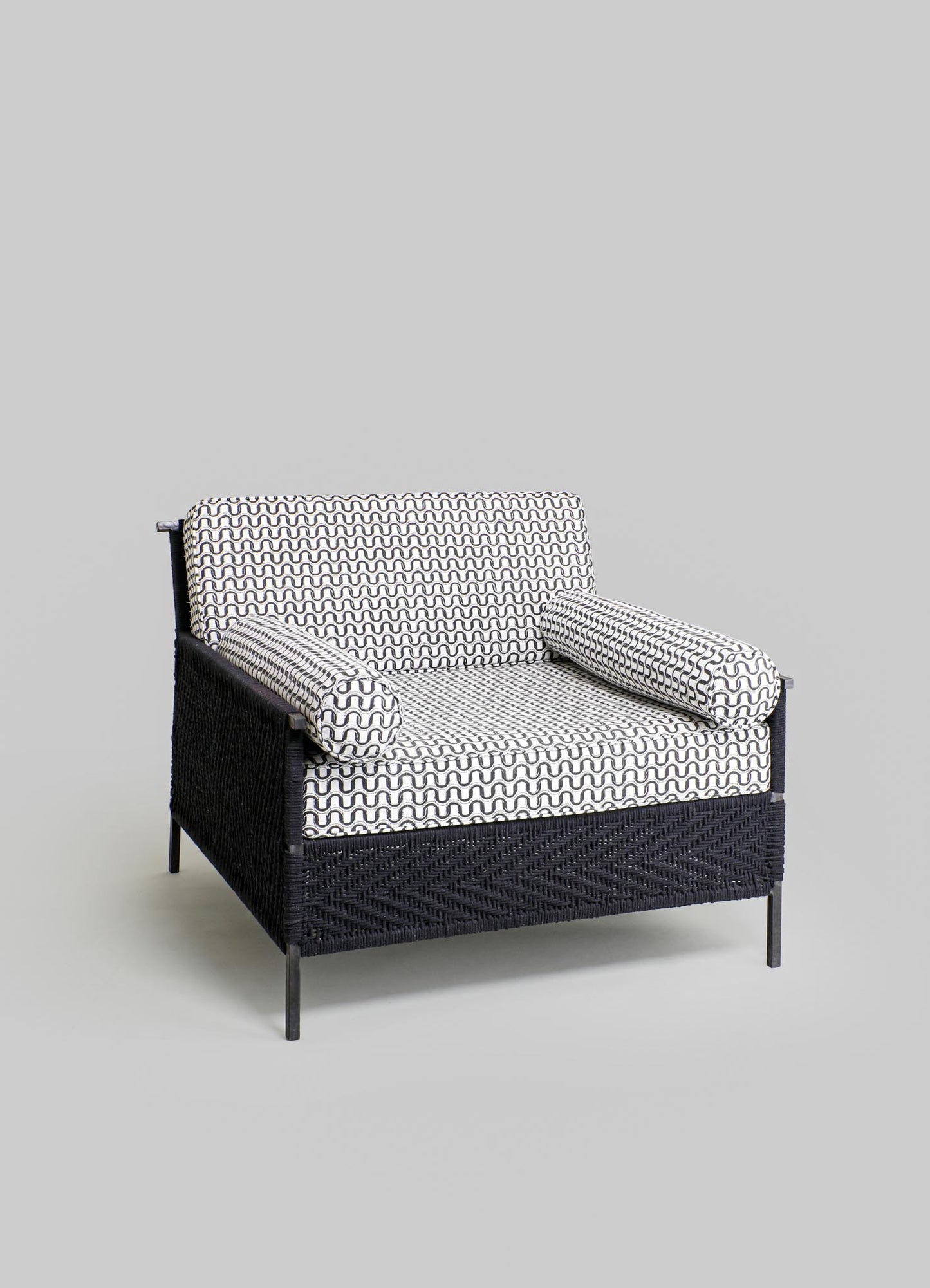 A black woven rattan loveseat with geometric pattern cushions in black and white fabric, featuring matching cylindrical bolster pillows, set against a light gray background.