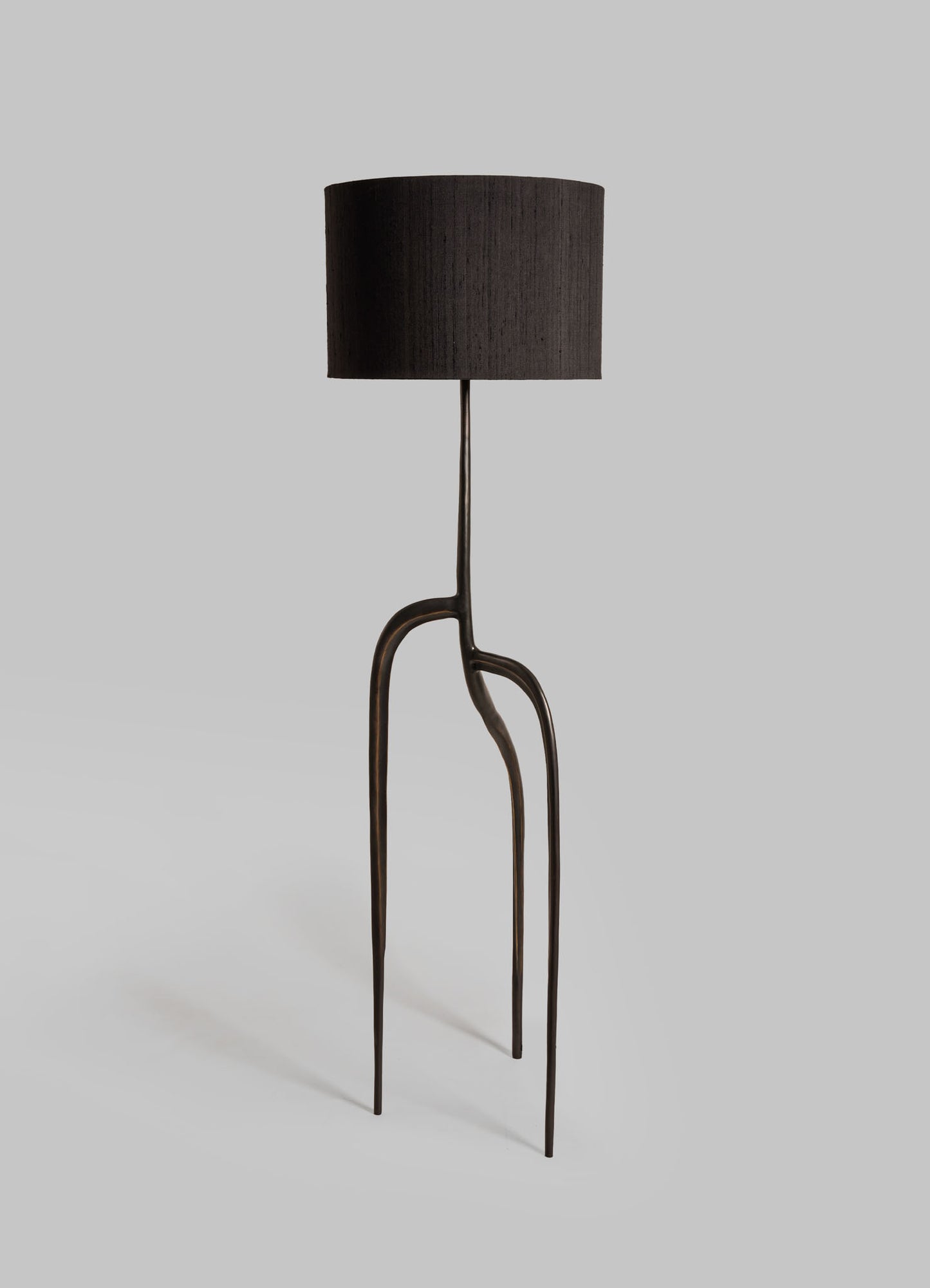 A floor lamp with a black fabric drum shade, featuring a sculptural metal base with organic, curving lines and a dark bronze finish.