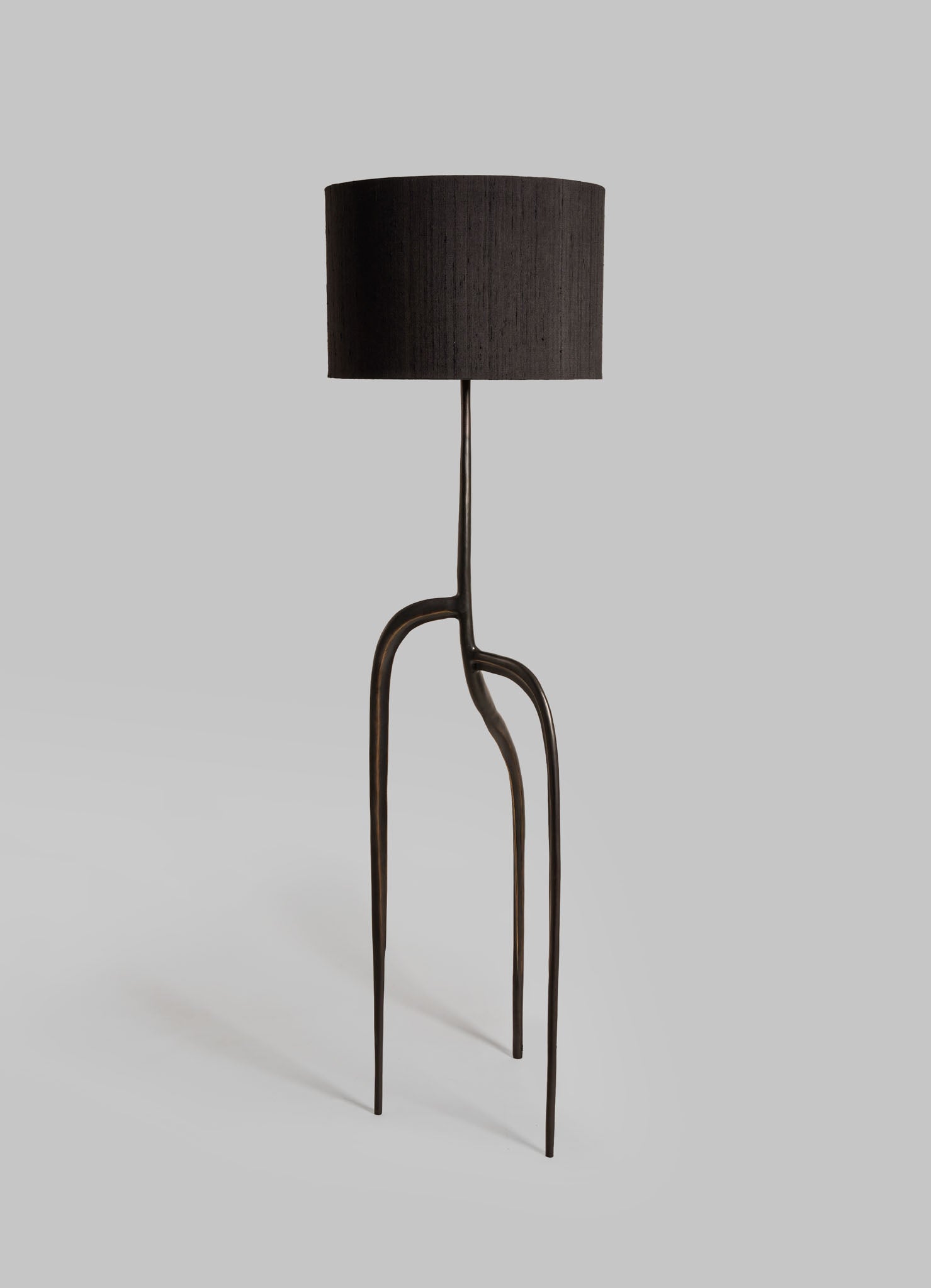 A floor lamp with a black fabric drum shade, featuring a sculptural metal base with organic, curving lines and a dark bronze finish.