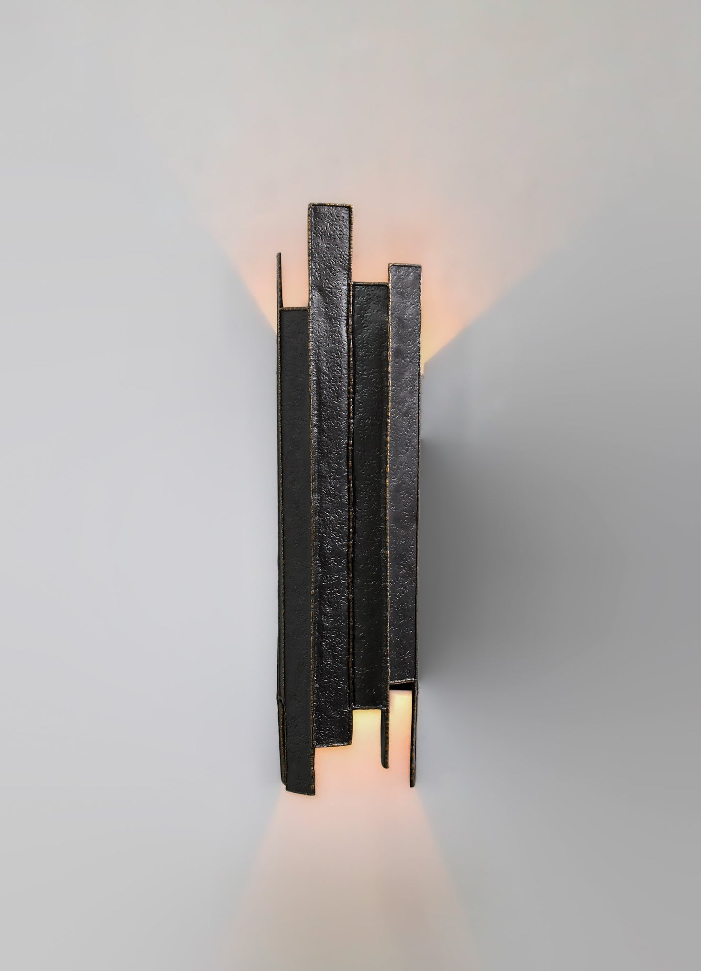 A wall-mounted light fixture with a modern, minimalist design featuring vertical black metal strips with a textured finish, emitting warm light upwards and downwards.