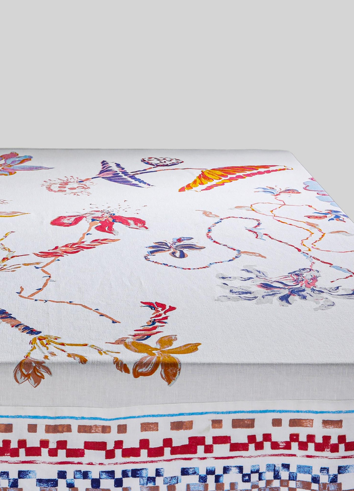 Colorful floral and geometric printed tablecloth on a white fabric base, featuring vibrant hues of red, orange, purple, and blue with a decorative border design. The material appears to be cotton or a similar textile.
