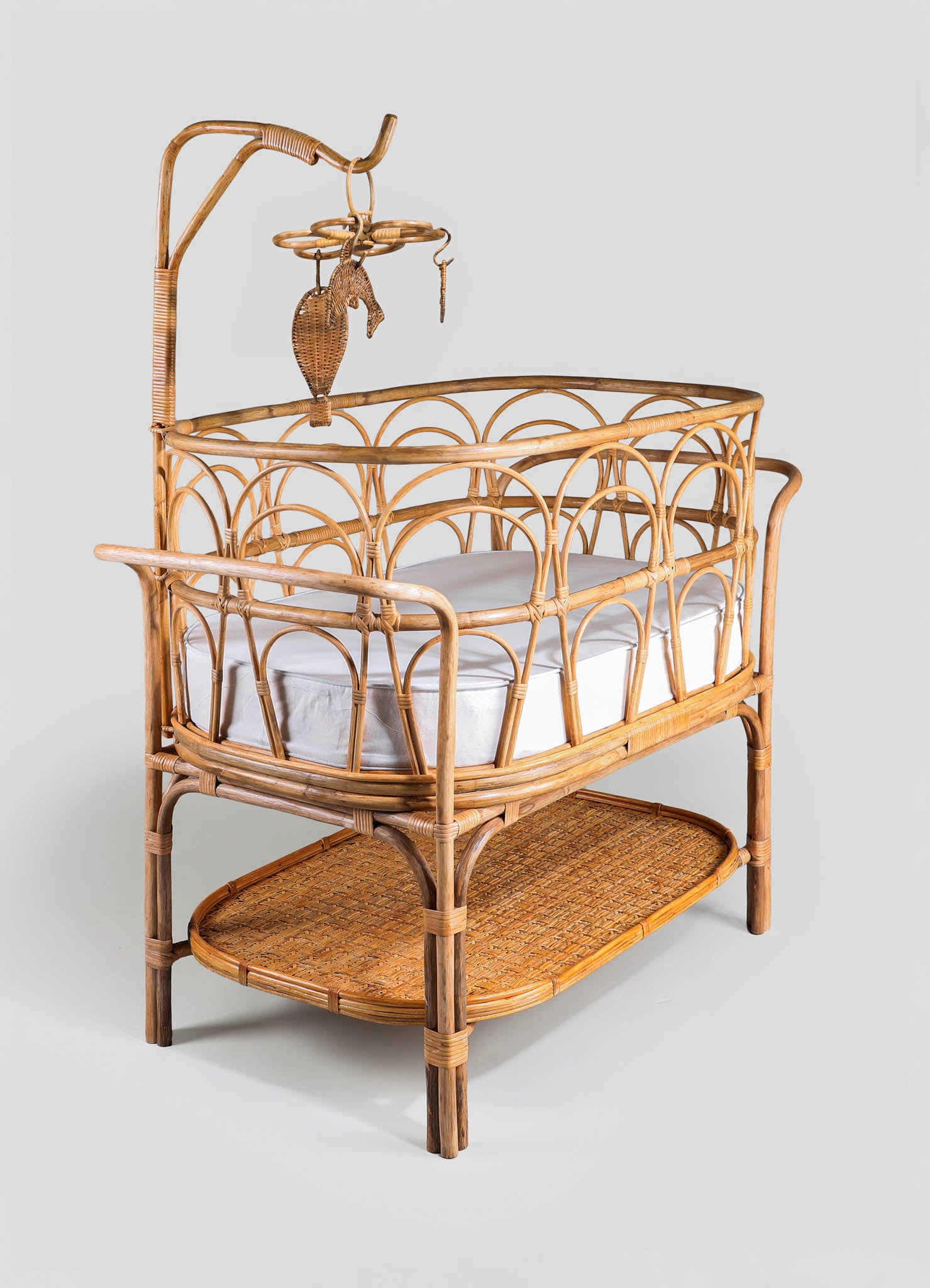 A rattan bassinet with a woven wicker frame, featuring a curved design with vertical and semi-circular patterns, supported by a lower rattan shelf. The bassinet has a white fabric-lined interior and an adjustable rattan canopy with hanging decorative elements.