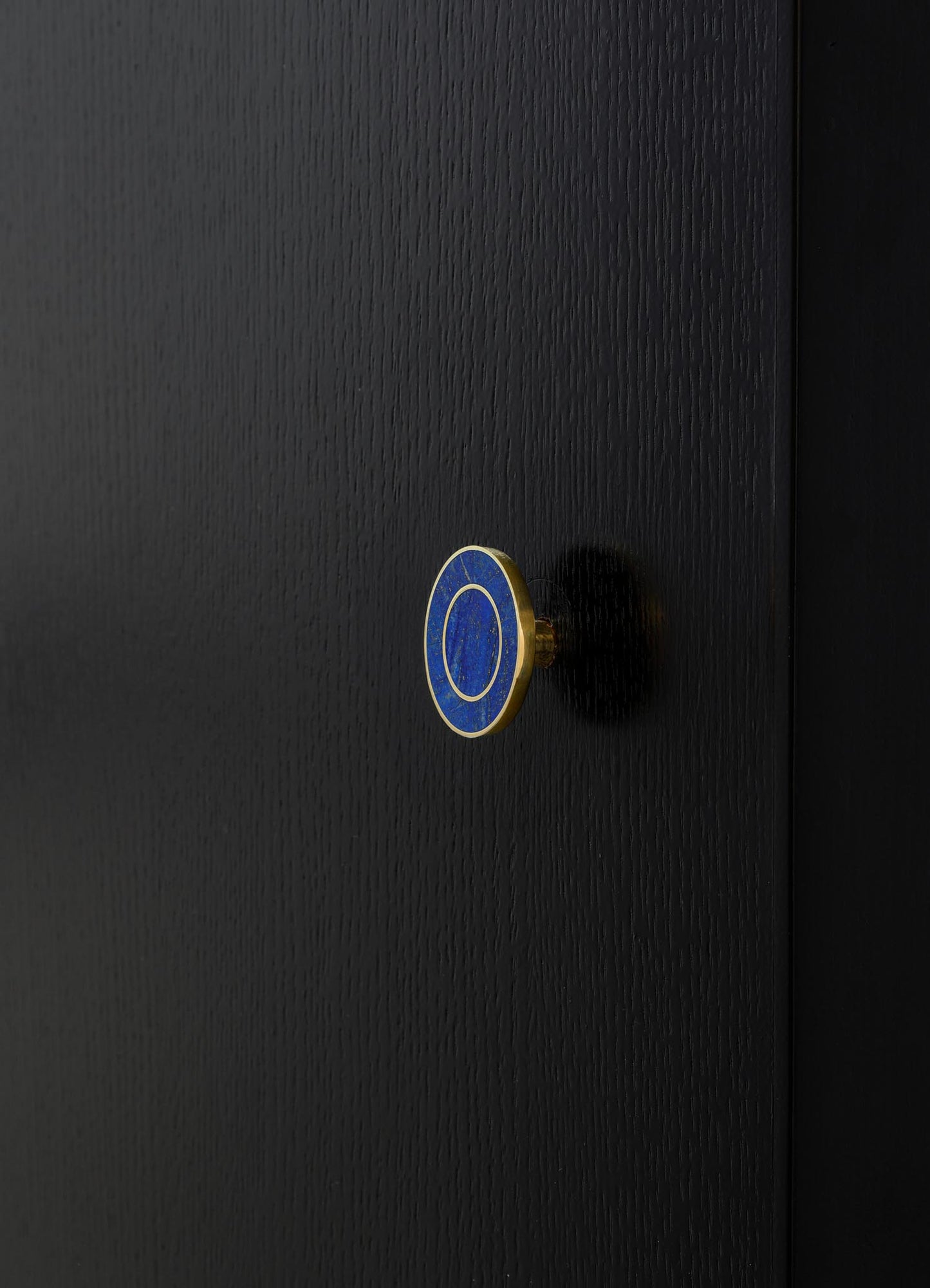 A close-up of a black textured surface with a circular brass and blue enameled knob, likely a piece of furniture or cabinetry hardware with a minimalist design.