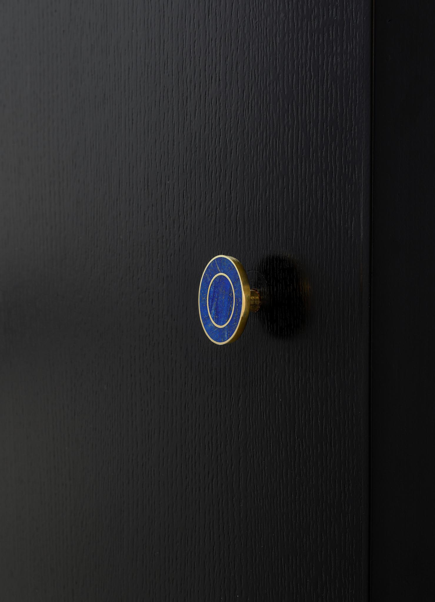 A close-up of a black textured surface with a circular brass and blue enameled knob, likely a piece of furniture or cabinetry hardware with a minimalist design.