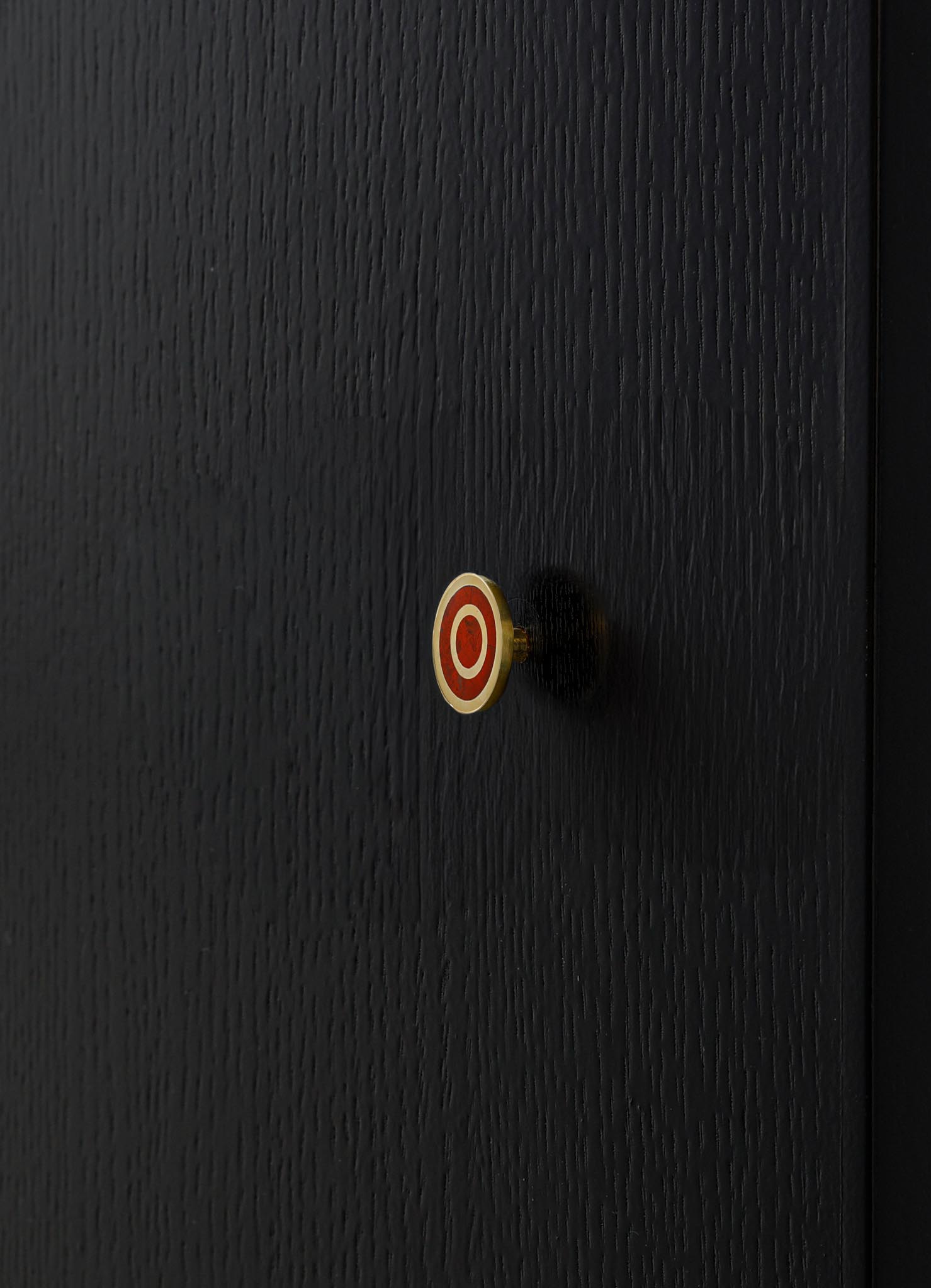 A black wooden surface with a textured finish features a small, round decorative knob with red and white concentric circles, casting a shadow on the wood.