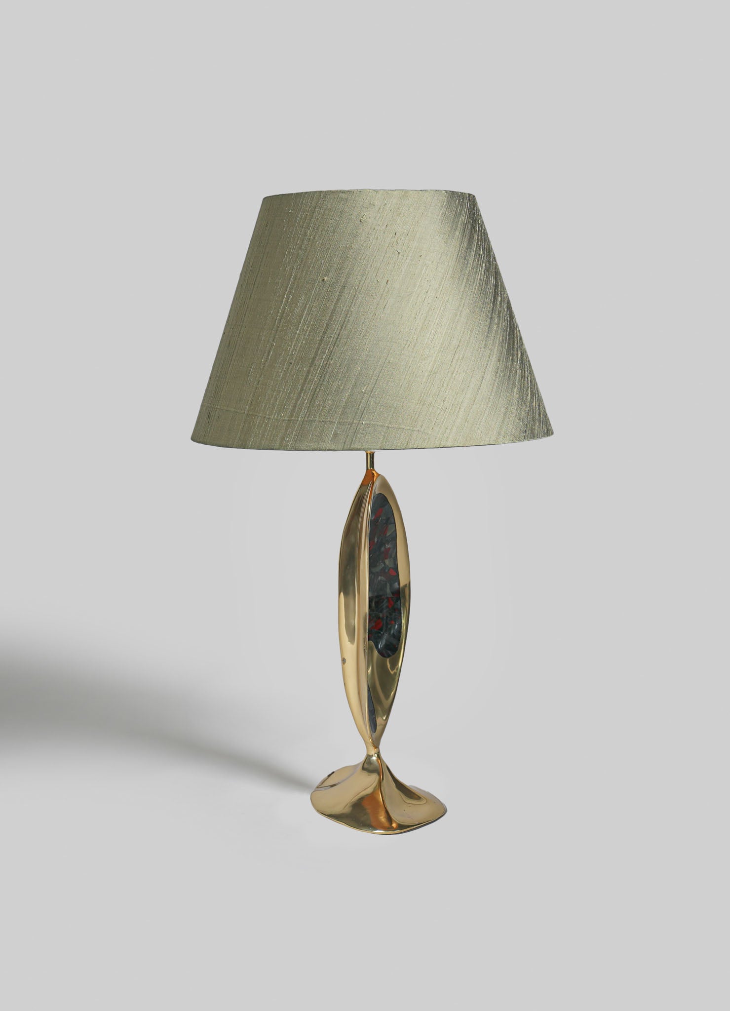 Table lamp with a brass base featuring an elongated oval shape and an abstract painted interior, topped with a textured beige fabric lampshade in a contemporary style.