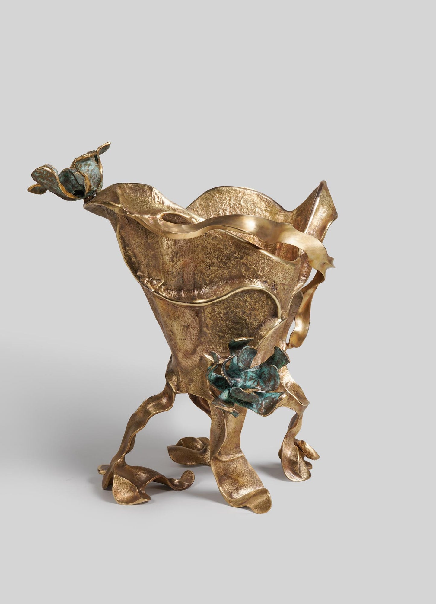 A decorative sculpture made of metal with a gold-tone finish, featuring abstract, flowing shapes and textured surfaces. It includes accents of patina in blue-green tones, and has an organic, sculptural form reminiscent of a floral or natural motif.