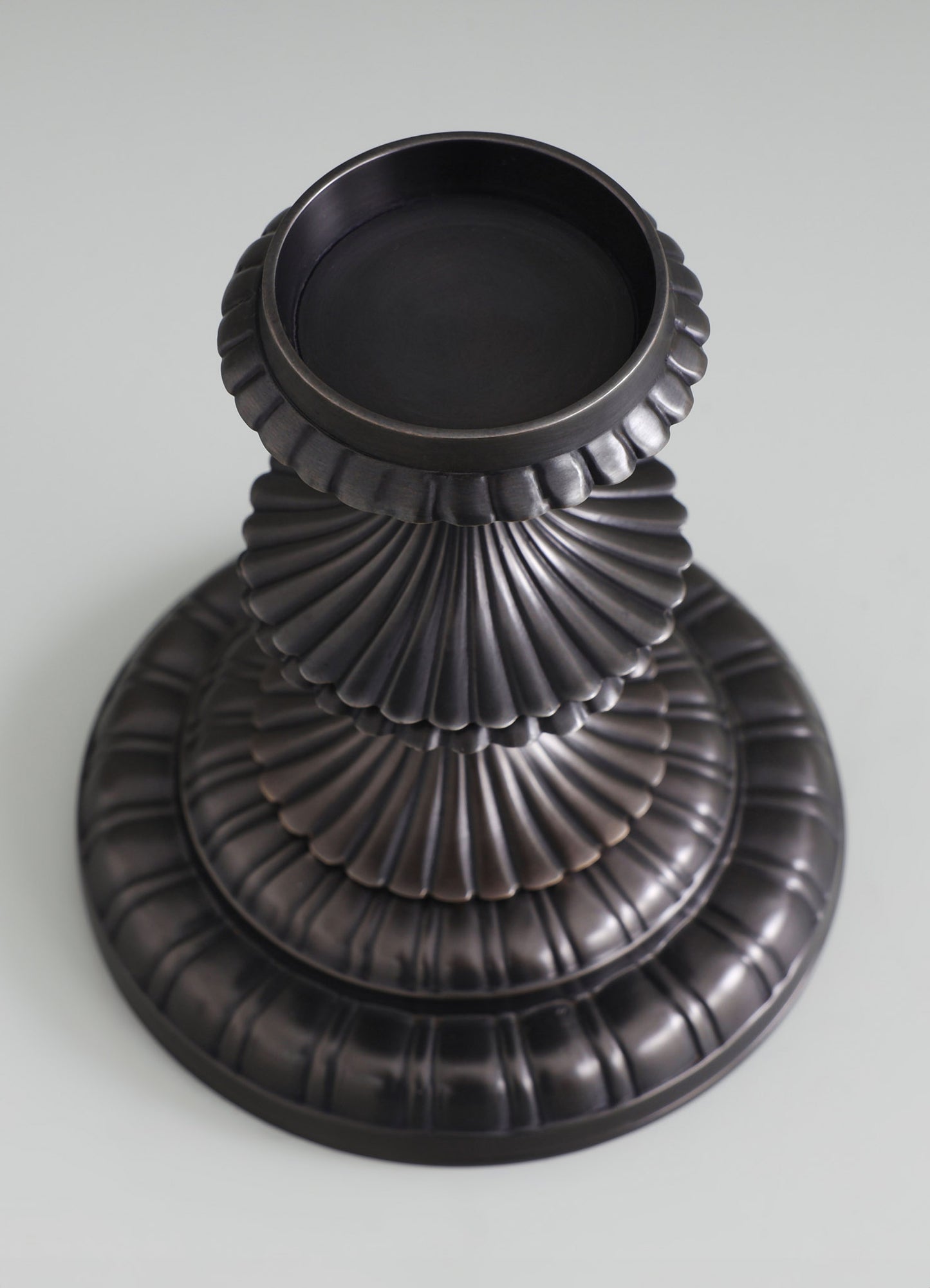 A black metal vase with a fluted, scalloped design featuring concentric ridges, set against a light background. The vase has a wide, circular opening and a textured surface with tiered, rounded edges.