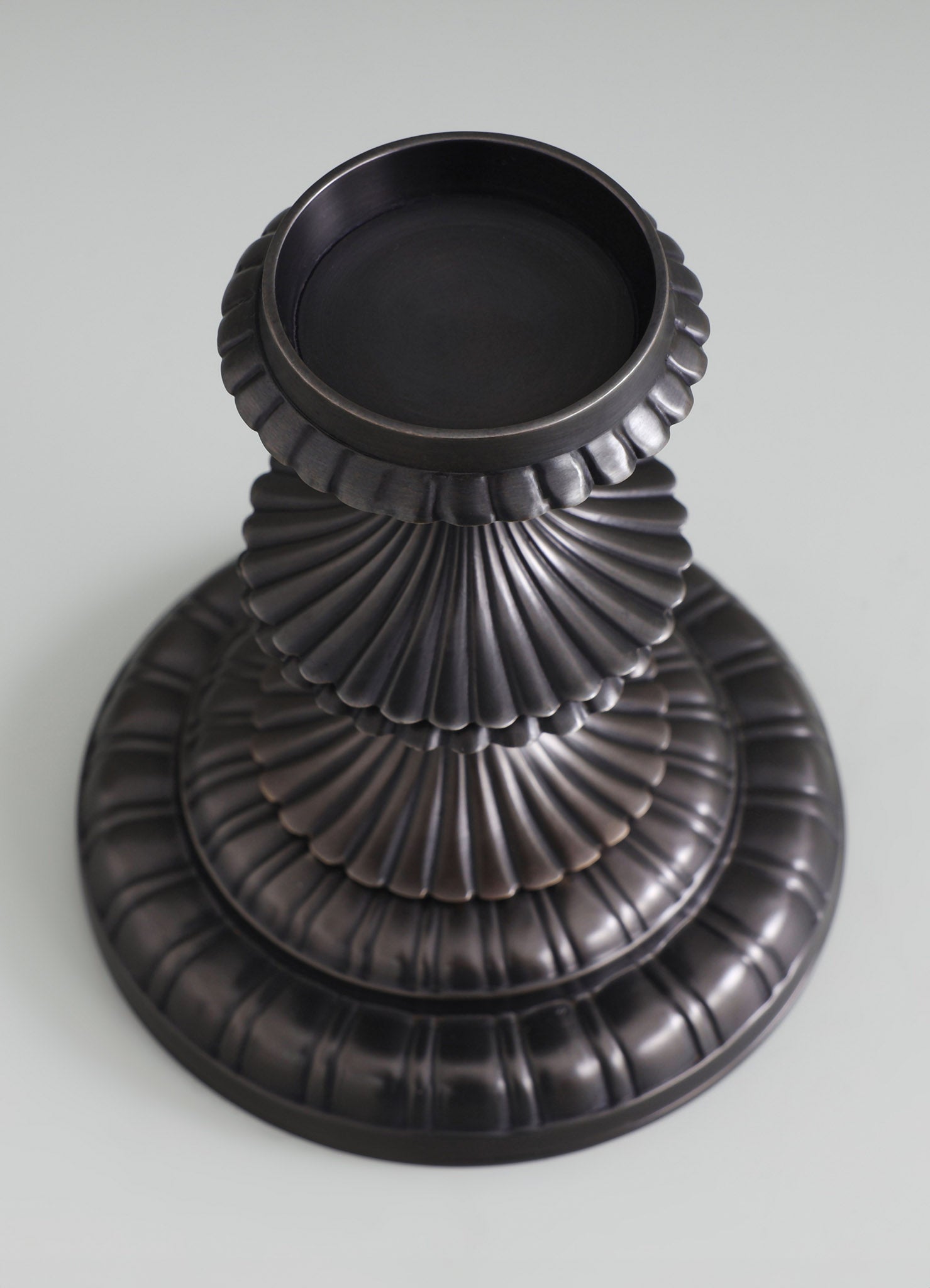 A black metal vase with a fluted, scalloped design featuring concentric ridges, set against a light background. The vase has a wide, circular opening and a textured surface with tiered, rounded edges.