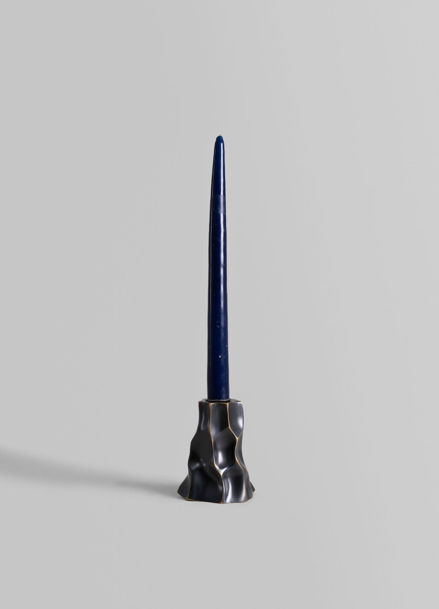 A tall, slender candle in deep blue wax is displayed in a sculptural brass holder with an irregular, organic shape and a dark, matte finish.