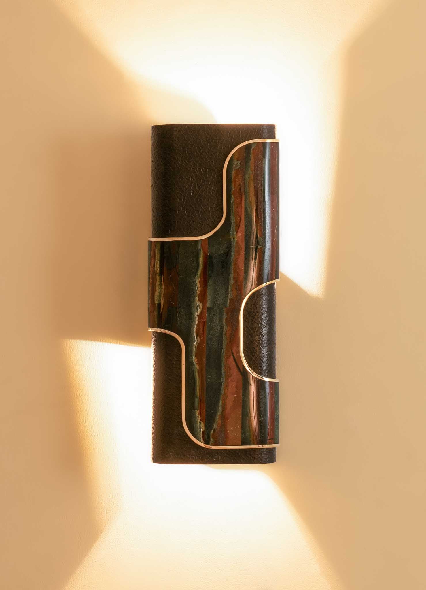 A cylindrical wall-mounted light fixture featuring a combination of textured black material and colorful, abstract marbled pattern with metallic gold accents, emitting warm light.