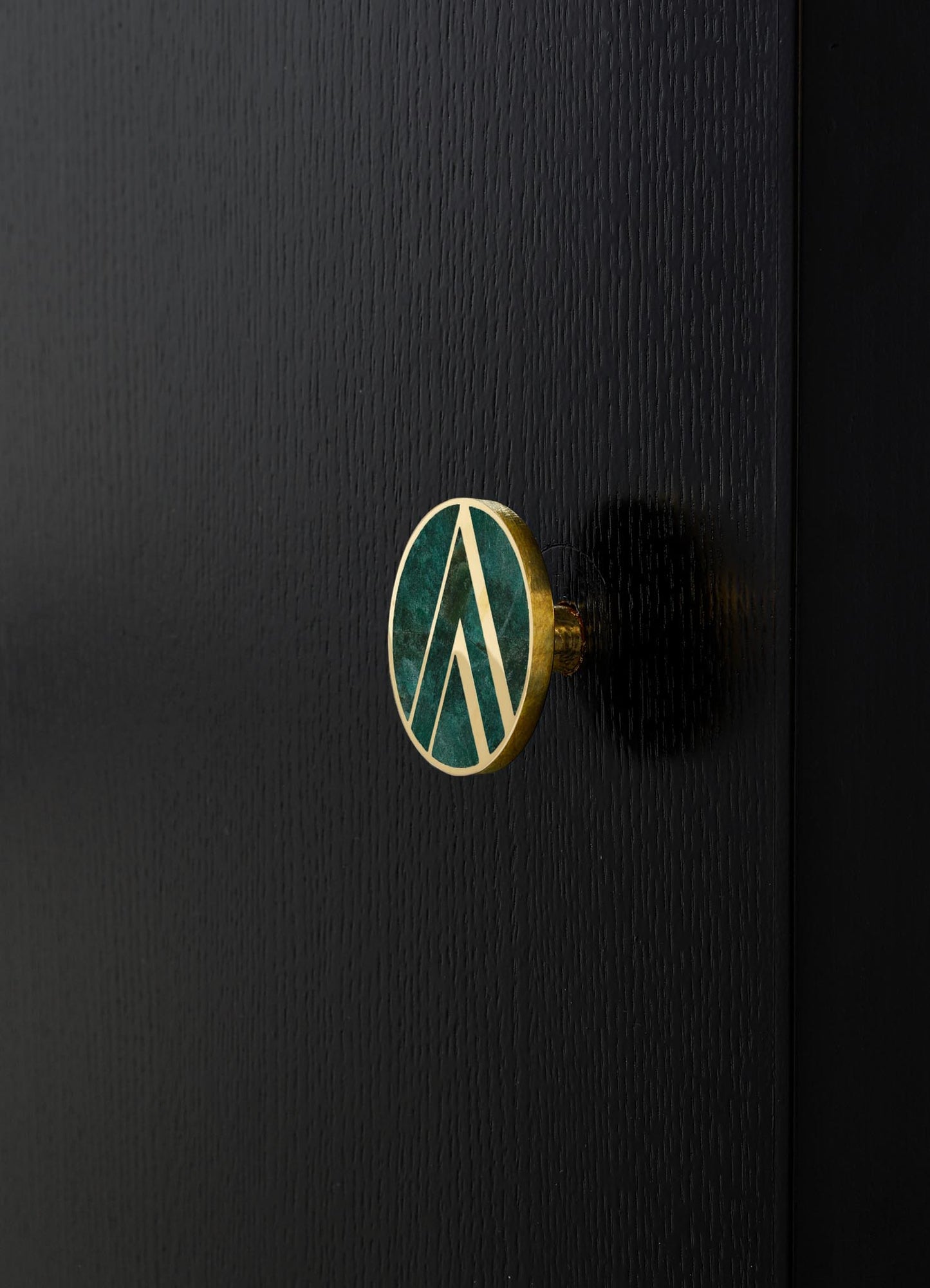 A round, decorative drawer knob with a green marble-like finish, accented by a gold metal border and geometric pattern, mounted on a black wooden surface.