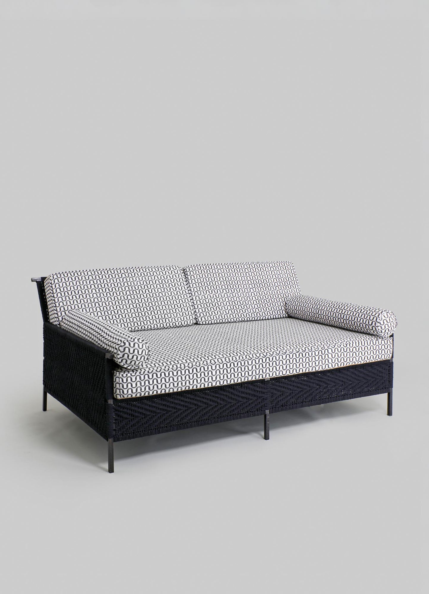 Woven black rattan sofa with cushions upholstered in a black and white geometric pattern, featuring cylindrical armrests and a sleek metal frame in a contemporary style.