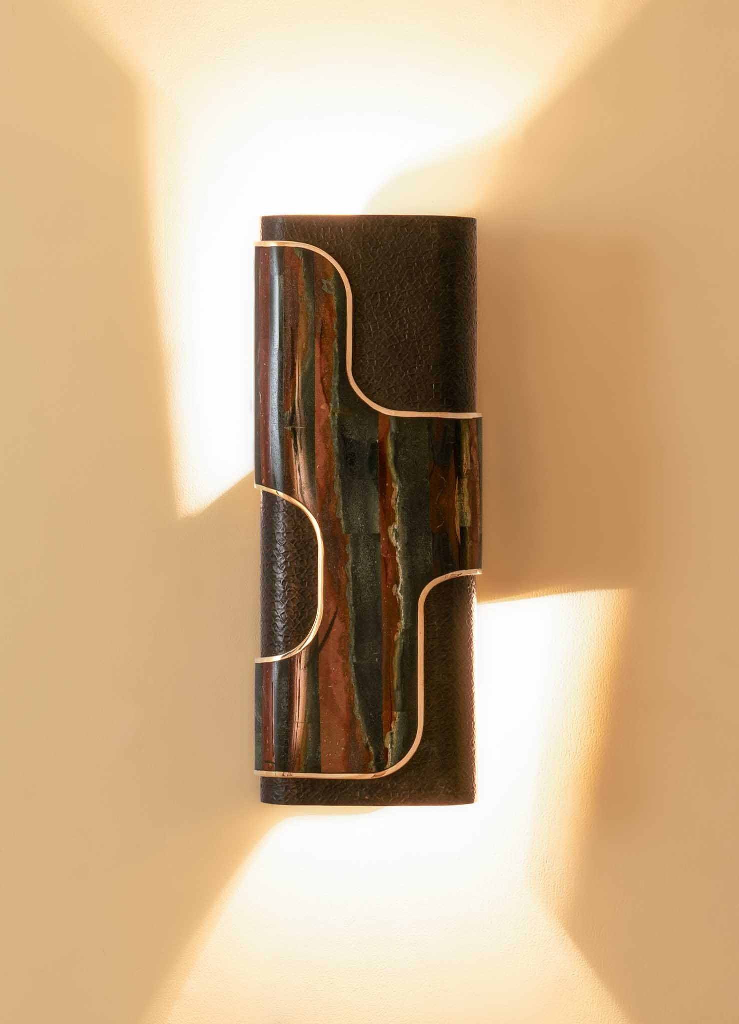 Wall-mounted light fixture with a cylindrical body made of textured dark material and an accent of multicolored marble with vertical streaks. The design features curved metallic accents and emits warm light onto a neutral-colored wall.