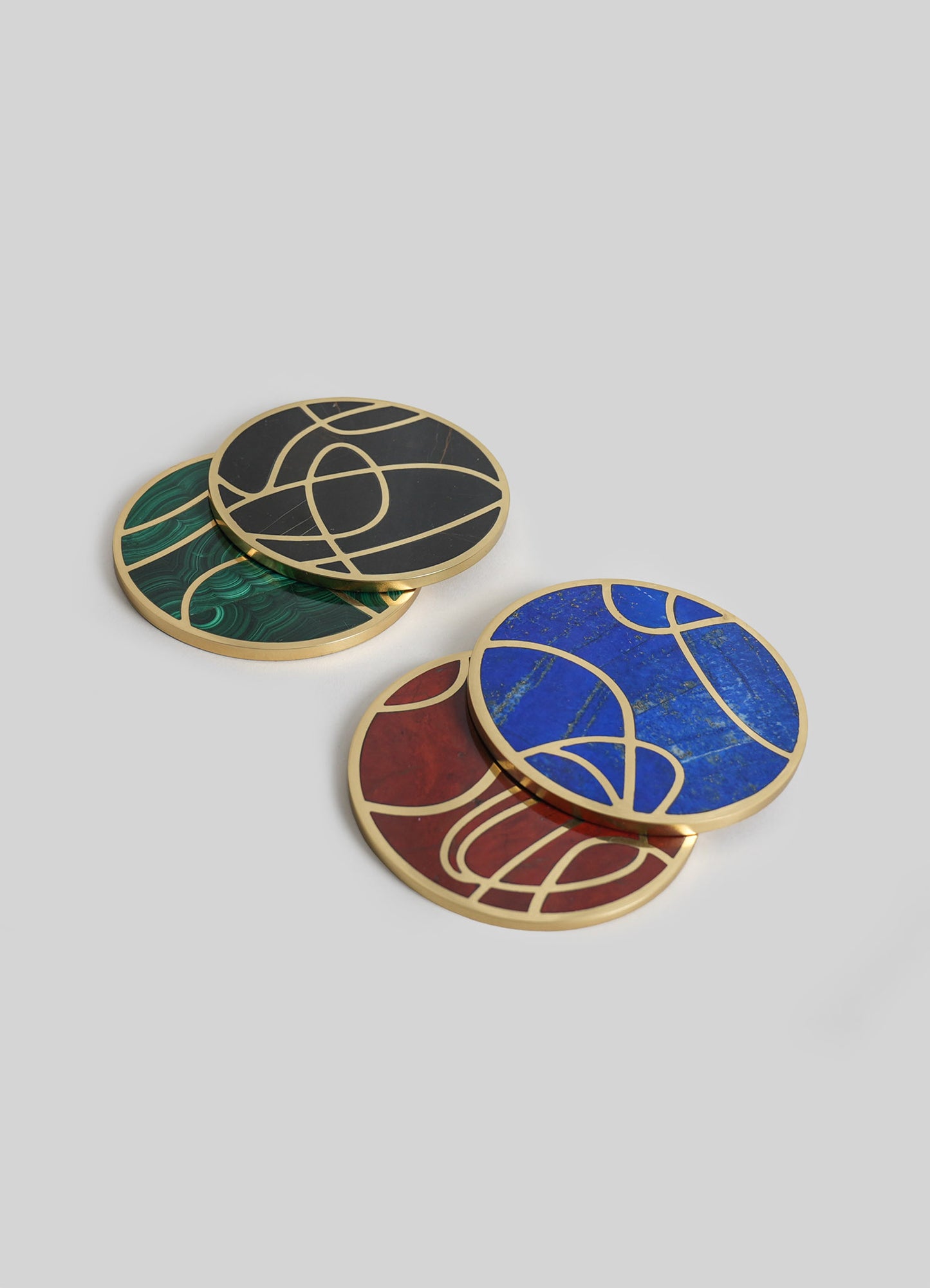 Set of four round coasters with abstract, curved line motifs in various bold colors: black with gold, green with gold, blue with gold, and red with gold. Made of materials resembling marble and metal, with a gold-tone border and accents, in a contemporary style.