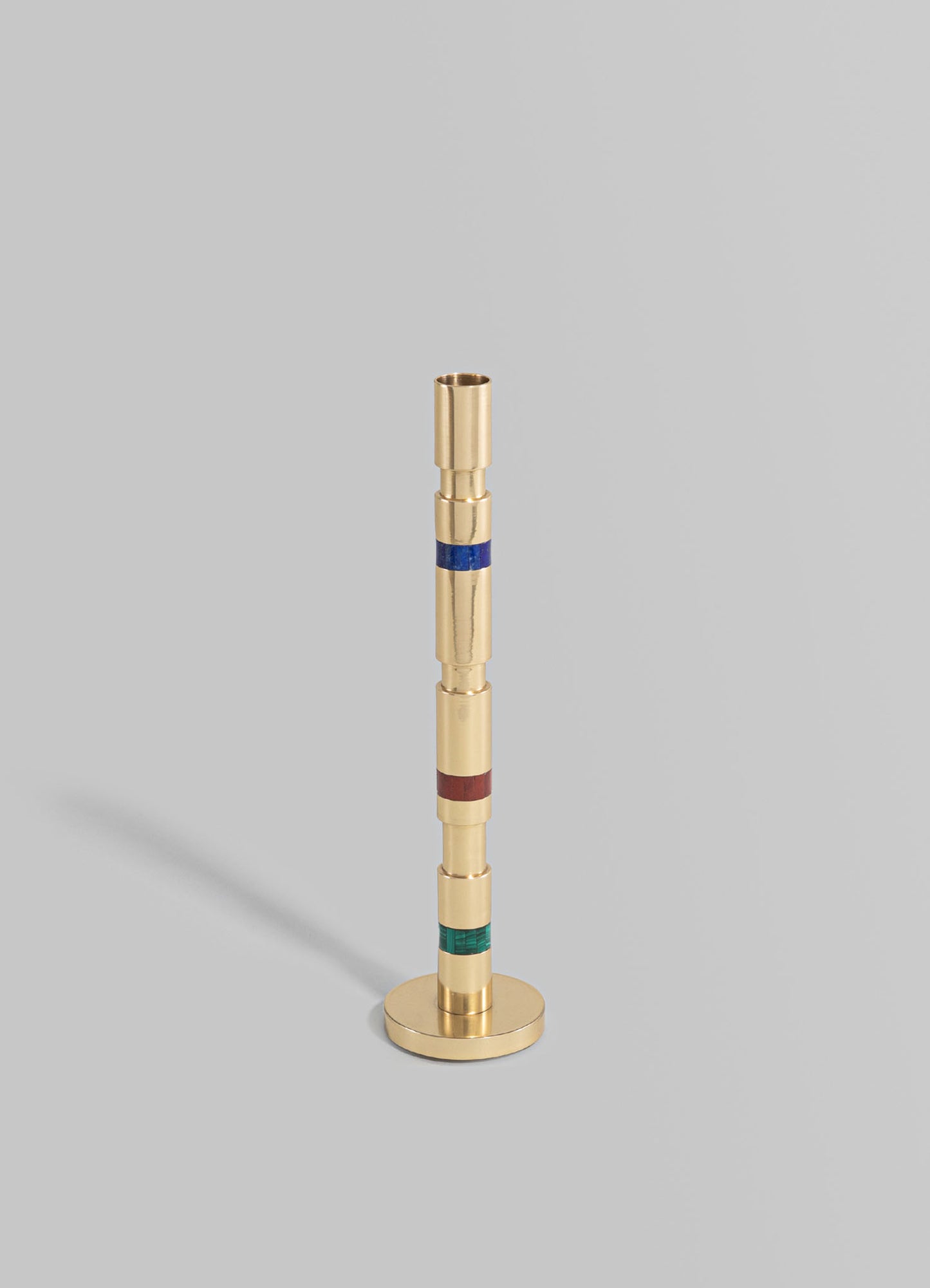 A brass candle holder with a tall, slender design featuring colorful striped accents in blue, red, and green, set on a circular base against a neutral background.