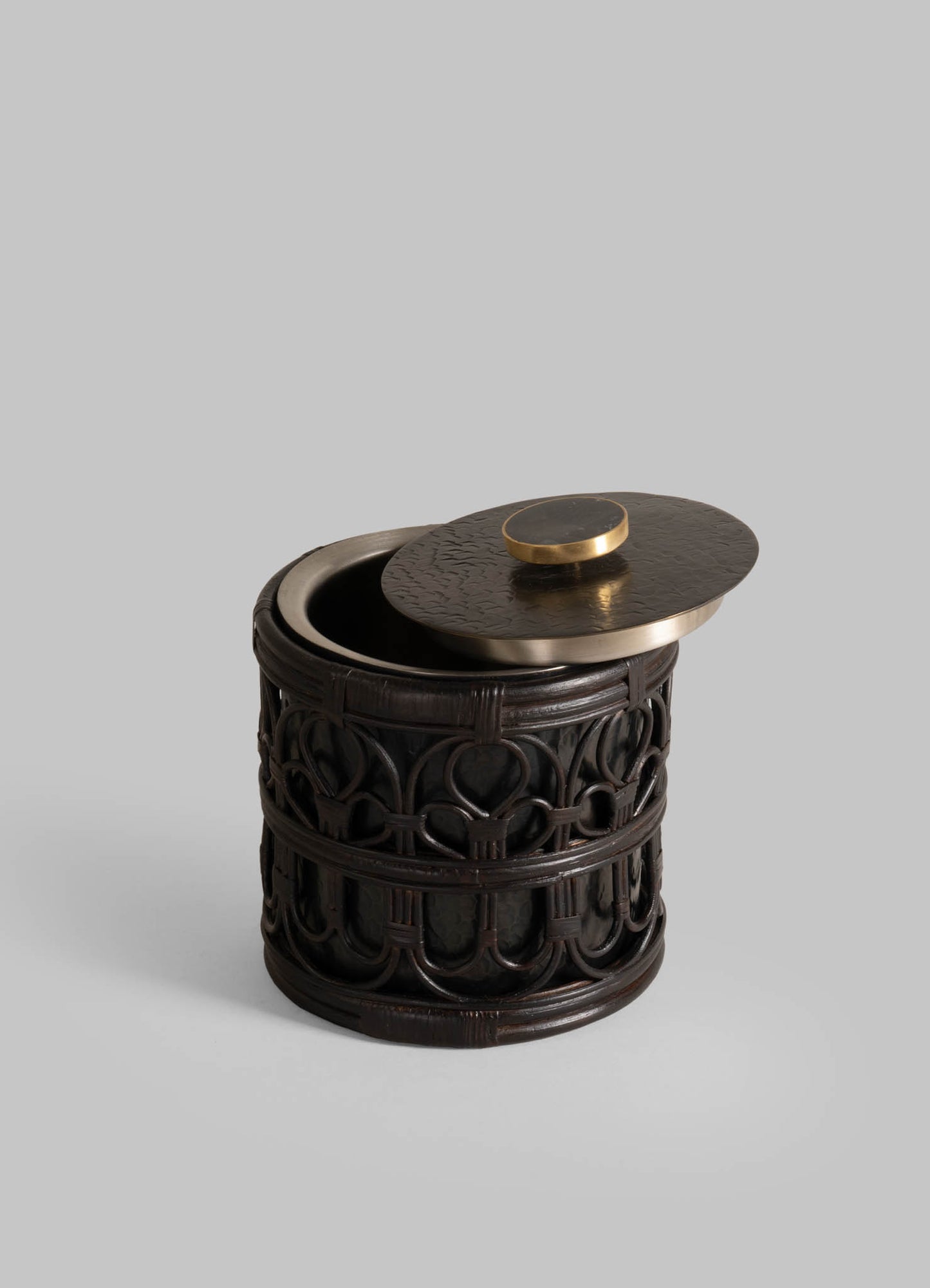 The VIYA Manipur Ice Bucket Small is a round, dark ornate container with intricate carvings and a hand-beaten brass lid with a circular handle, shown partially open against a light gray background.