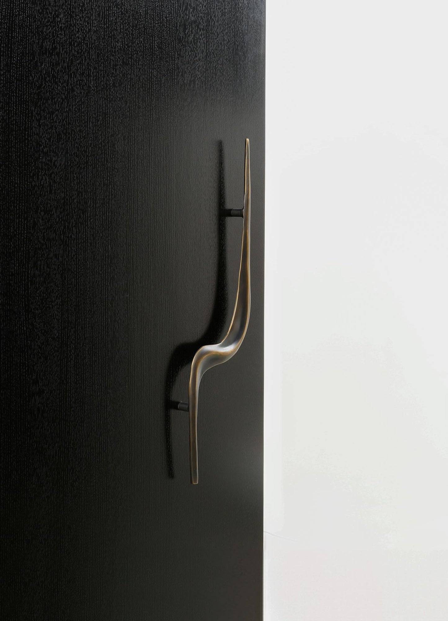 A modern wall hook made of metal with a sleek, curved design, featuring a metallic finish mounted on a black textured wall. The background includes a contrasting white surface, emphasizing the contemporary style of the hook.
