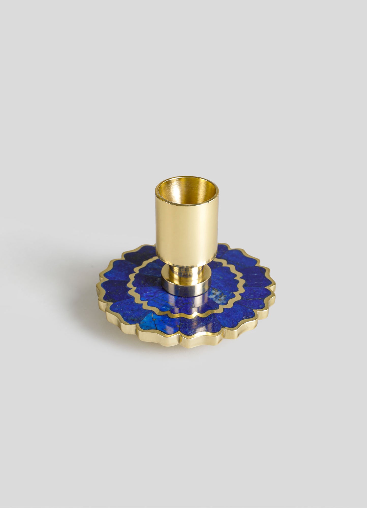 A gold-colored metal candle holder with a cylindrical design sits on a decorative blue and gold scalloped ceramic plate with a floral motif. The combination of materials includes metal and glazed ceramic, featuring vibrant blue tones and metallic accents.