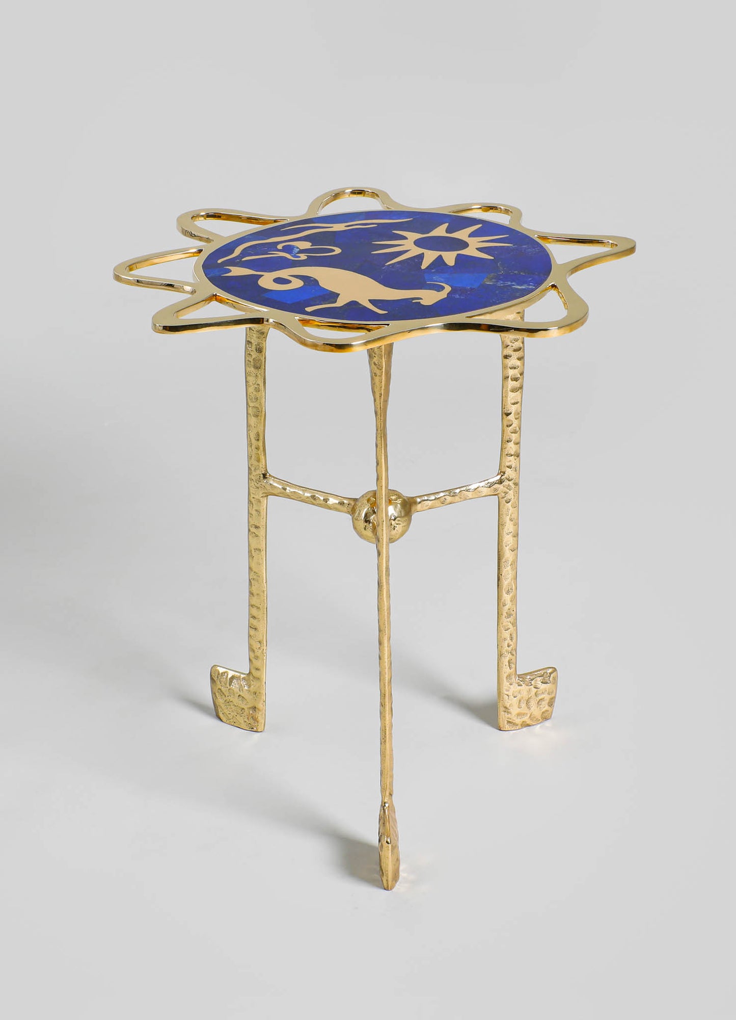 A small side table with a gold-toned, hammered metal base and three legs, topped with an oval-shaped glass surface featuring a decorative blue motif resembling a star and animal figures.
