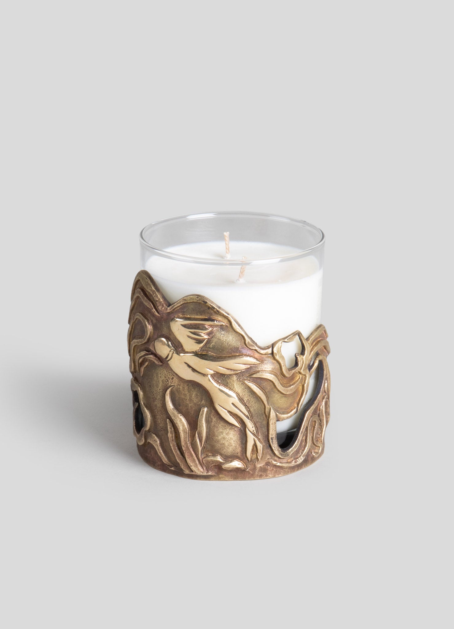 A glass candle holder with a decorative metal sleeve featuring an organic, flowing design in antique brass finish. The clear glass container holds a white pillar candle.
