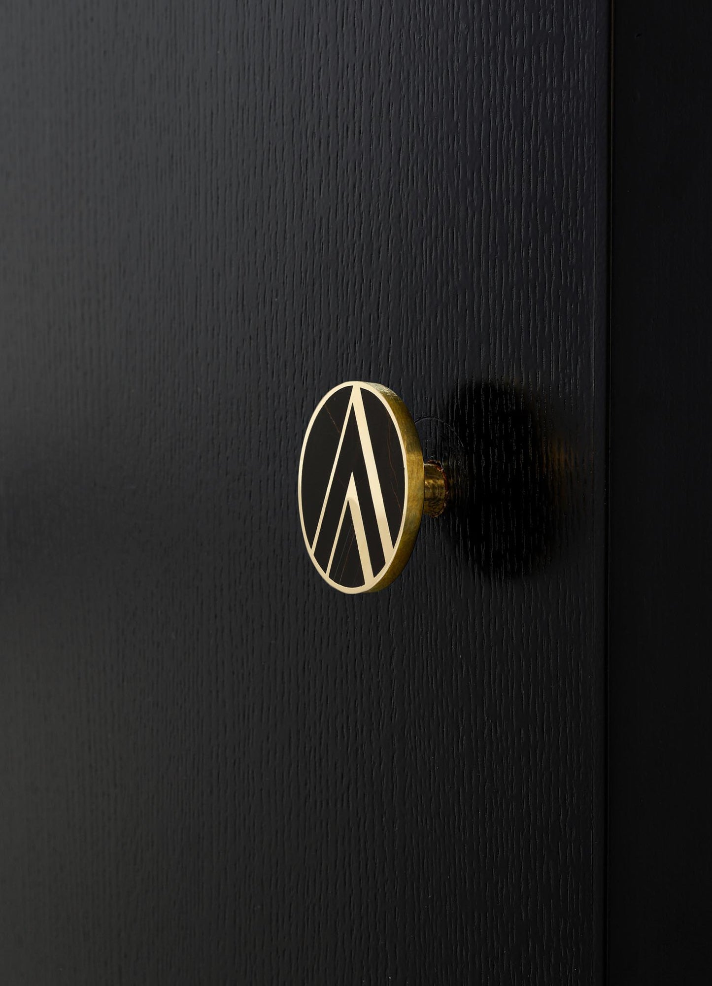 A black wooden door with a textural finish and a round brass handle featuring a geometric design in black and brass tones.
