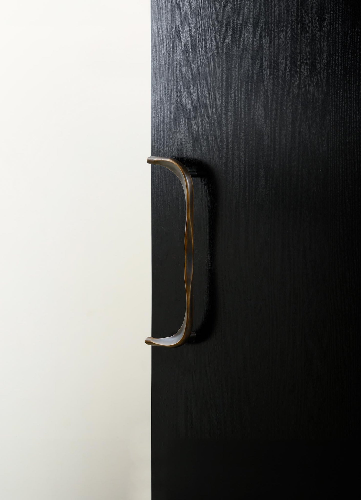 A close-up view of a black textured door with a sleek, curved metallic handle. The door features a minimalist design with a matte finish, contrasting against a light background.