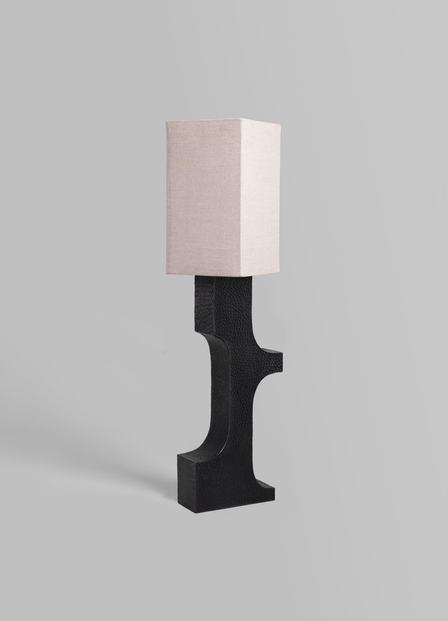A table lamp with a rectangular fabric shade in soft beige and a sculptural base in black textured material, exhibiting a modern and minimalist style.