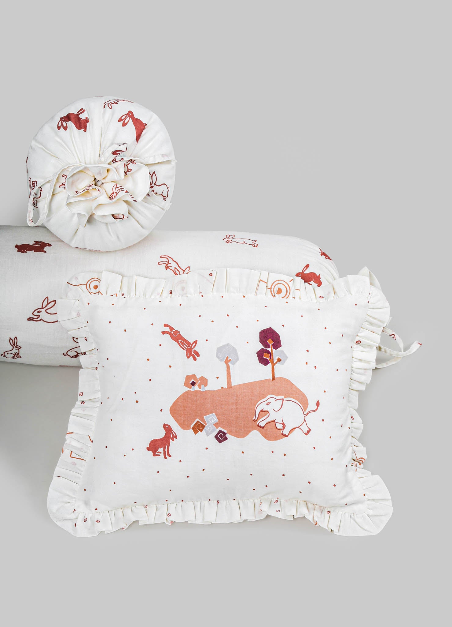 Textile accessories including a pillow and a ruffled cushion cover in white fabric with embroidered animal and nature motifs in shades of red, orange, and gray. The materials appear to be soft cotton or linen, featuring ruffled edges for a decorative, handcrafted style.