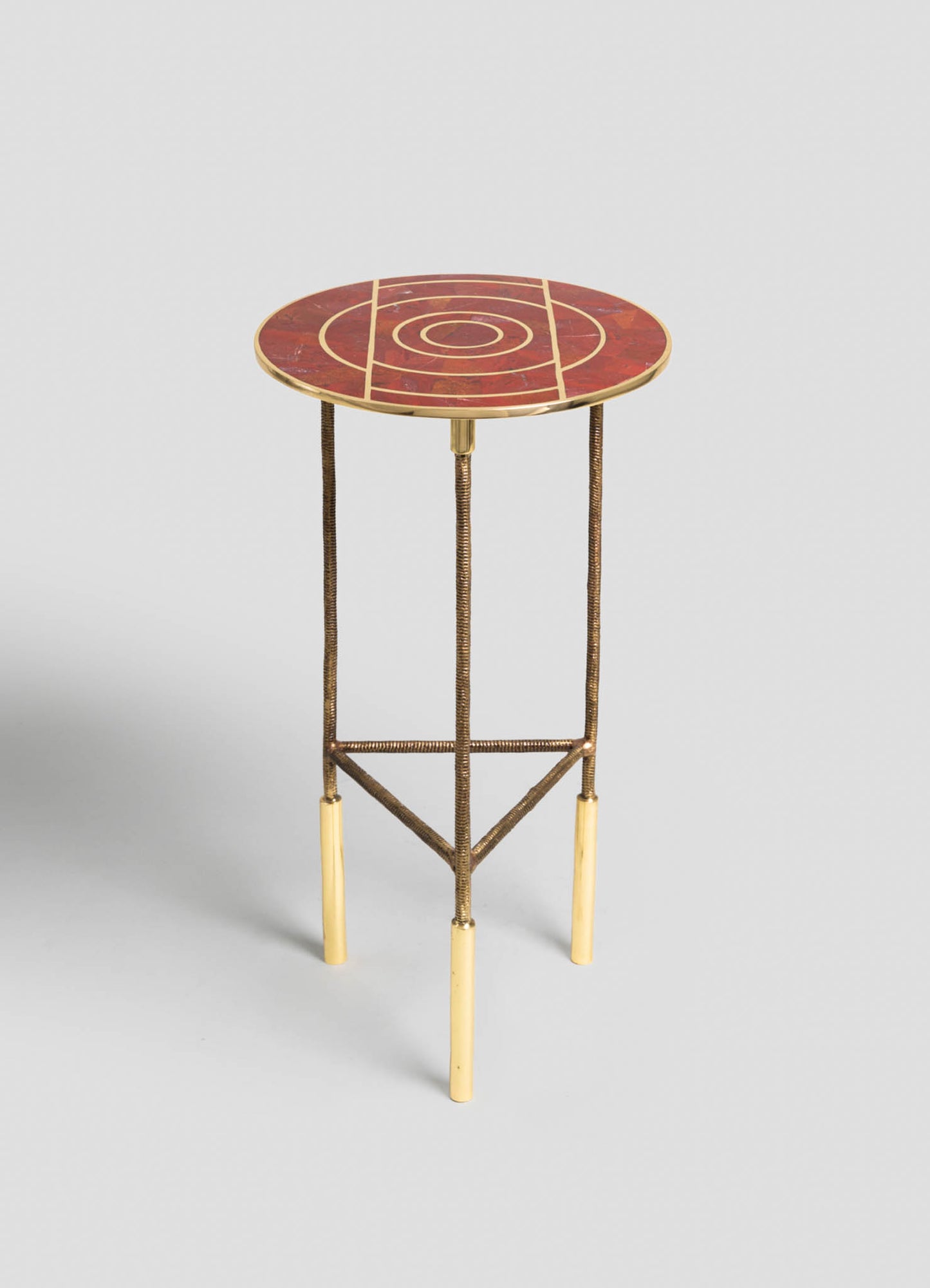A round side table with a patterned red and gold marble top featuring concentric circles and geometric lines, supported by slender metal legs with a brass finish and beige accents at the feet.