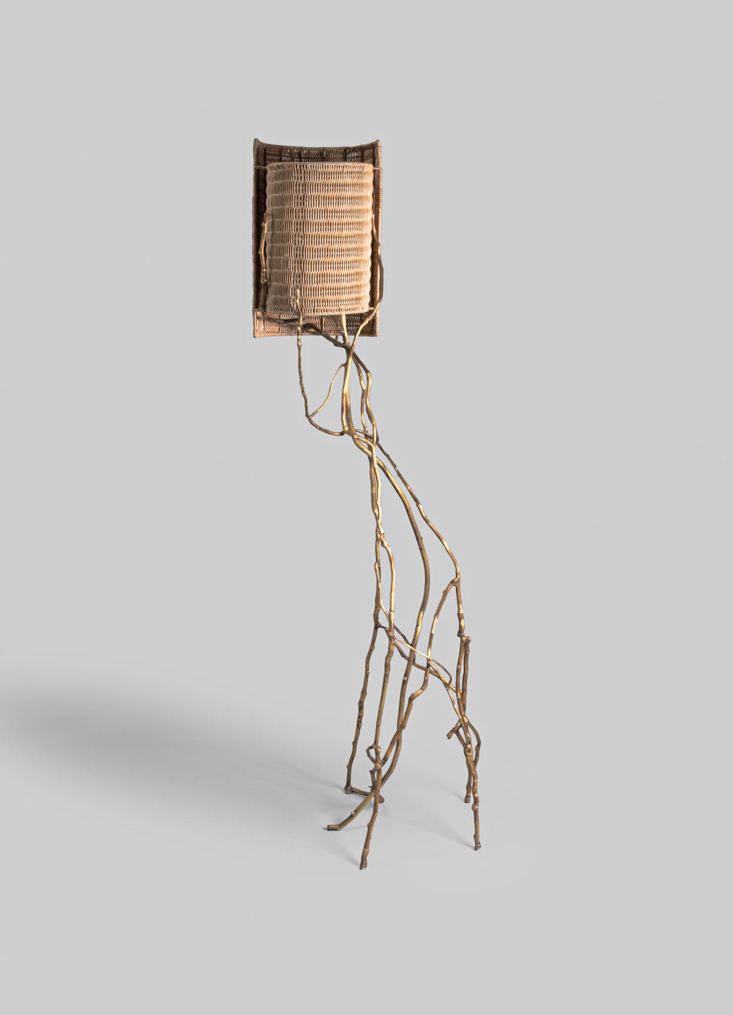 A tall floor lamp with a base made of intertwined, natural wood branches and a woven rattan lampshade in a neutral beige tone. The design is rustic and organic, emphasizing handcrafted materials and a natural aesthetic.