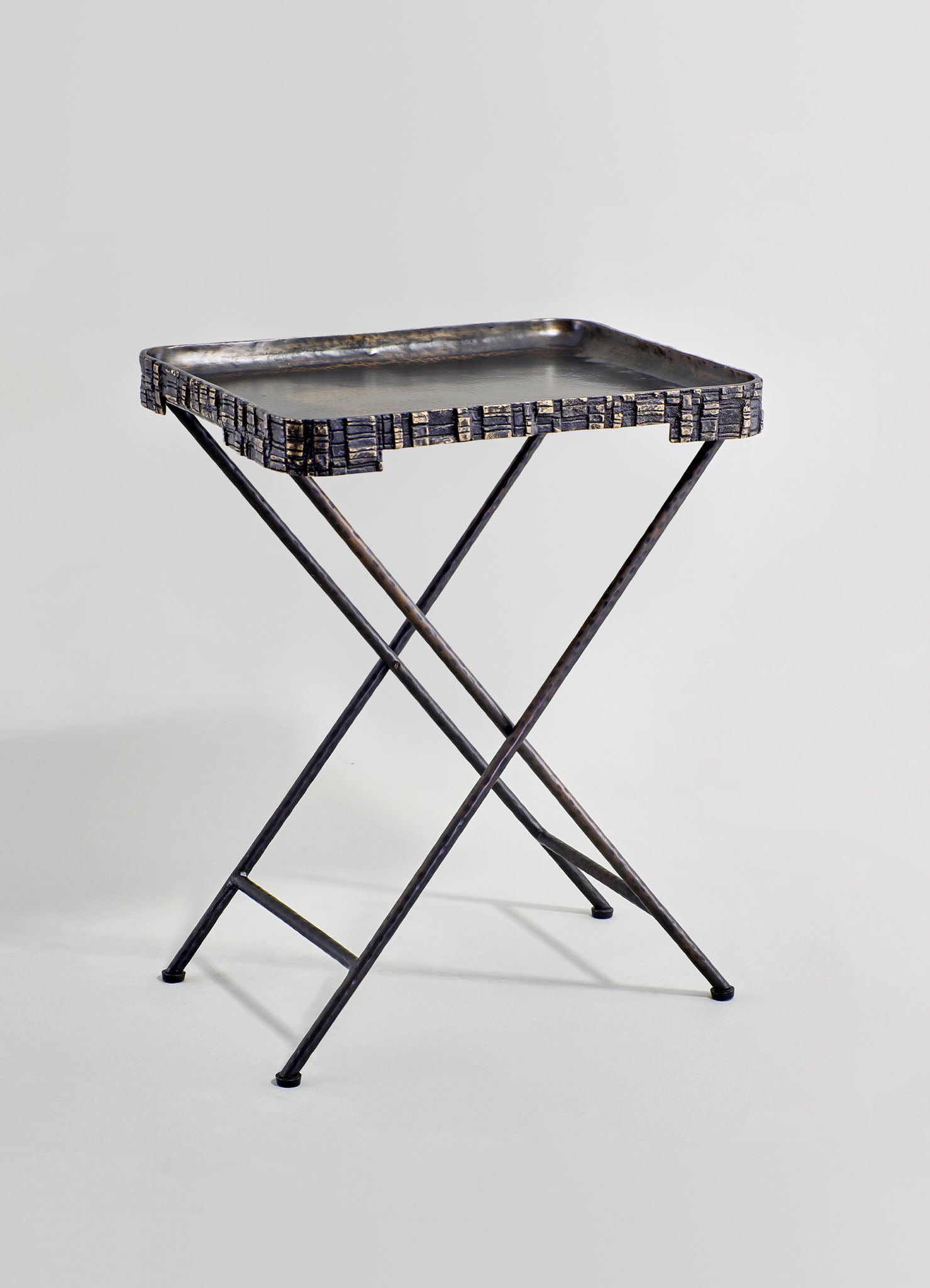 A small folding tray table with a rectangular metal tray top and crossed metal legs, featuring a dark, rustic finish. The tray has a textured, embossed border pattern and a smooth surface, giving it an industrial, vintage style.
