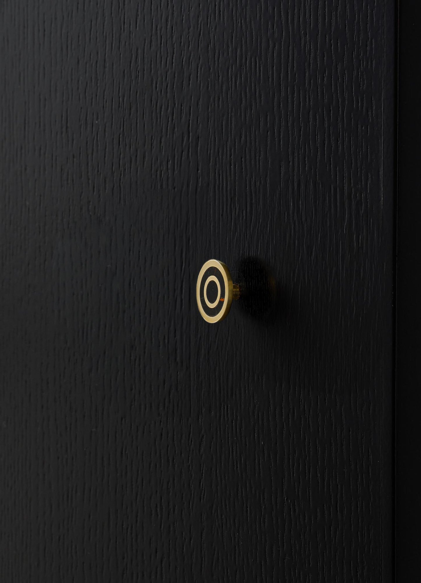 A black textured door featuring a small, circular brass doorknob with a concentric black and brass design element in the center. The surface has a vertical grain pattern, and the overall style appears modern and minimalist.