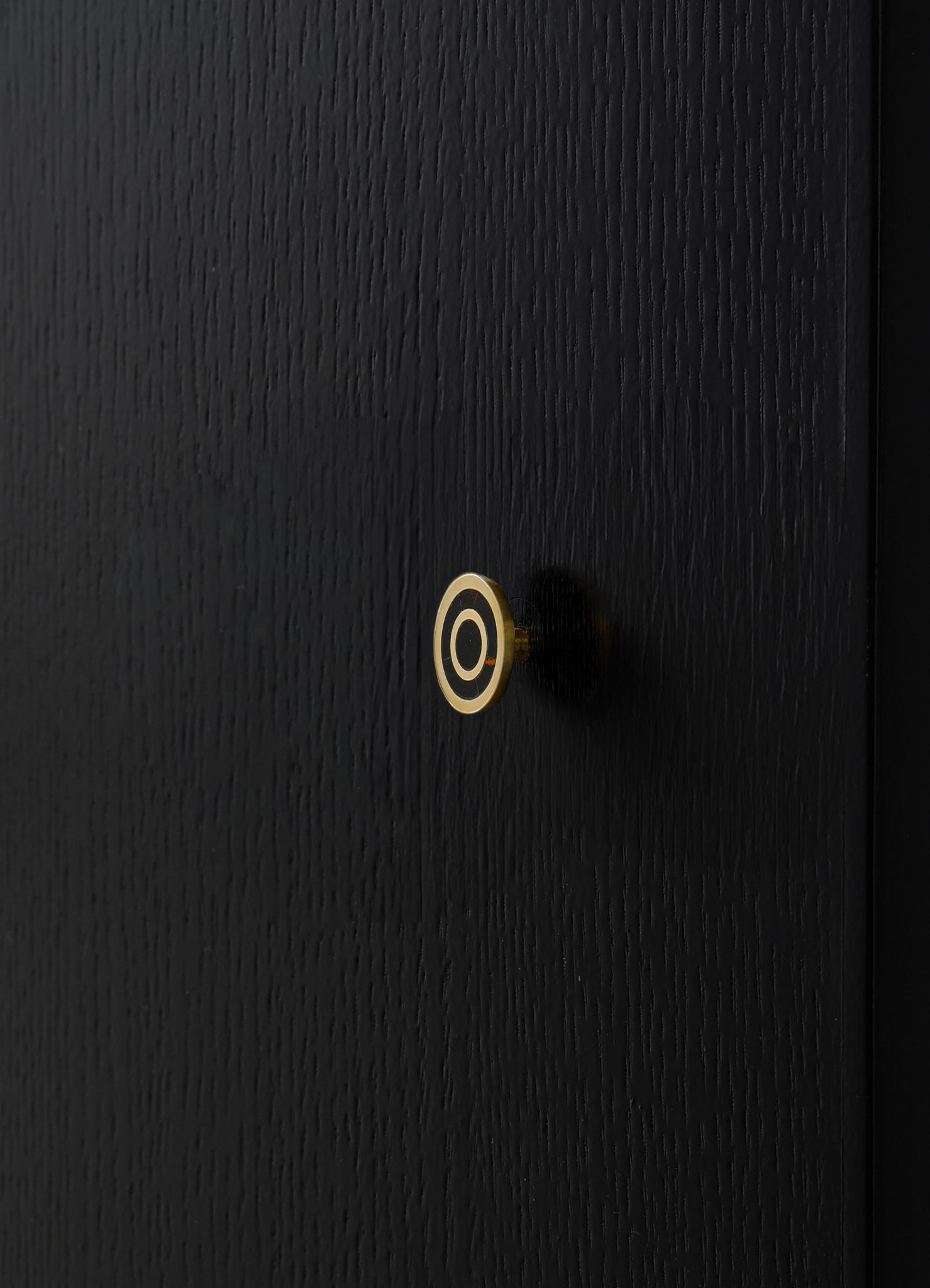 A black textured door featuring a small, circular brass doorknob with a concentric black and brass design element in the center. The surface has a vertical grain pattern, and the overall style appears modern and minimalist.