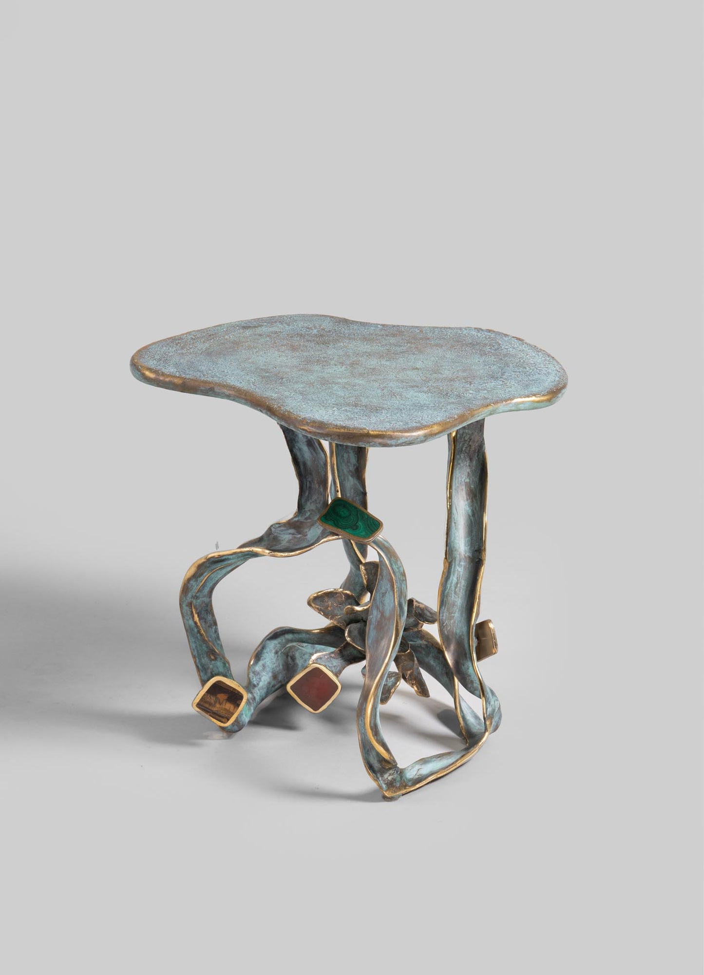 A small side table with a textured, irregularly shaped tabletop in a distressed metallic finish, featuring sculptural, curving legs with a patina effect. The legs include decorative square accents in gold and contrasting colors.